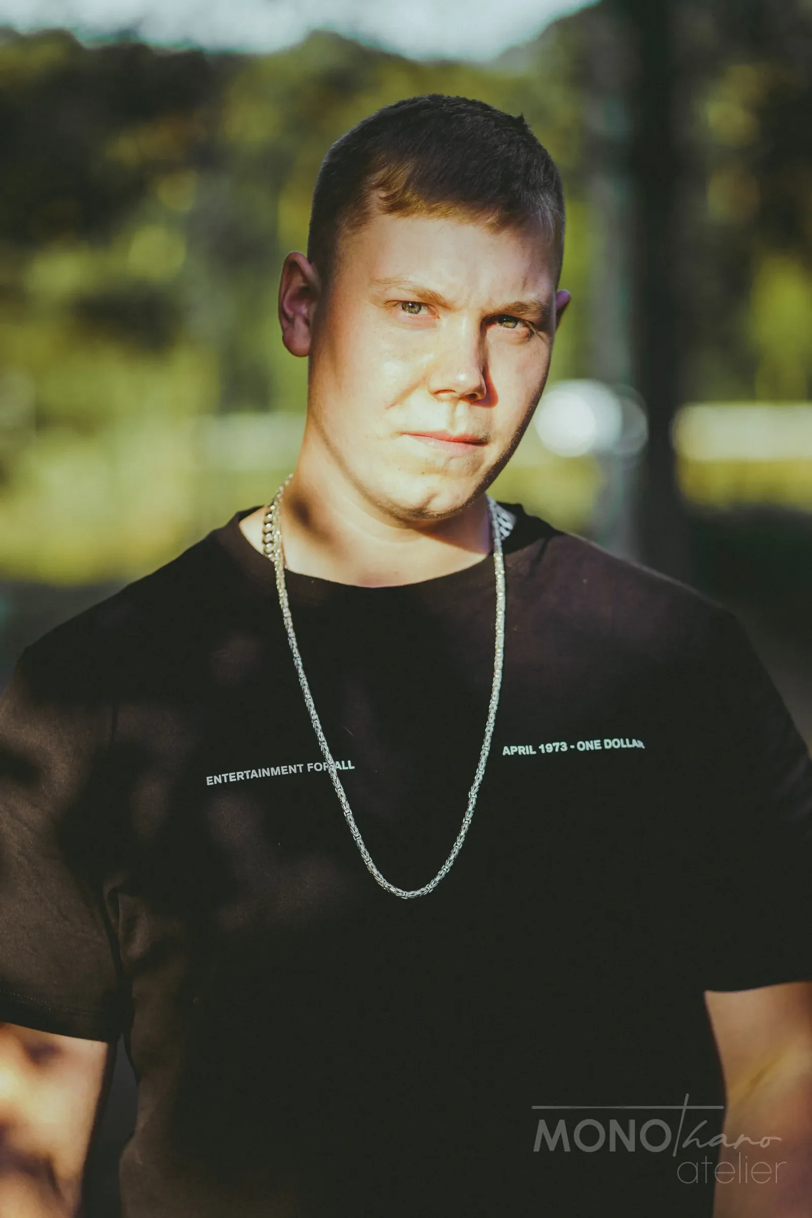 Nahaufnahme eines Mannes mit kurzen dunklen Haaren, schwarzem T-Shirt mit weißen Textstücken und silberner Kette, im Freien bei Sonnenlicht, unscharfer grüner Hintergrund