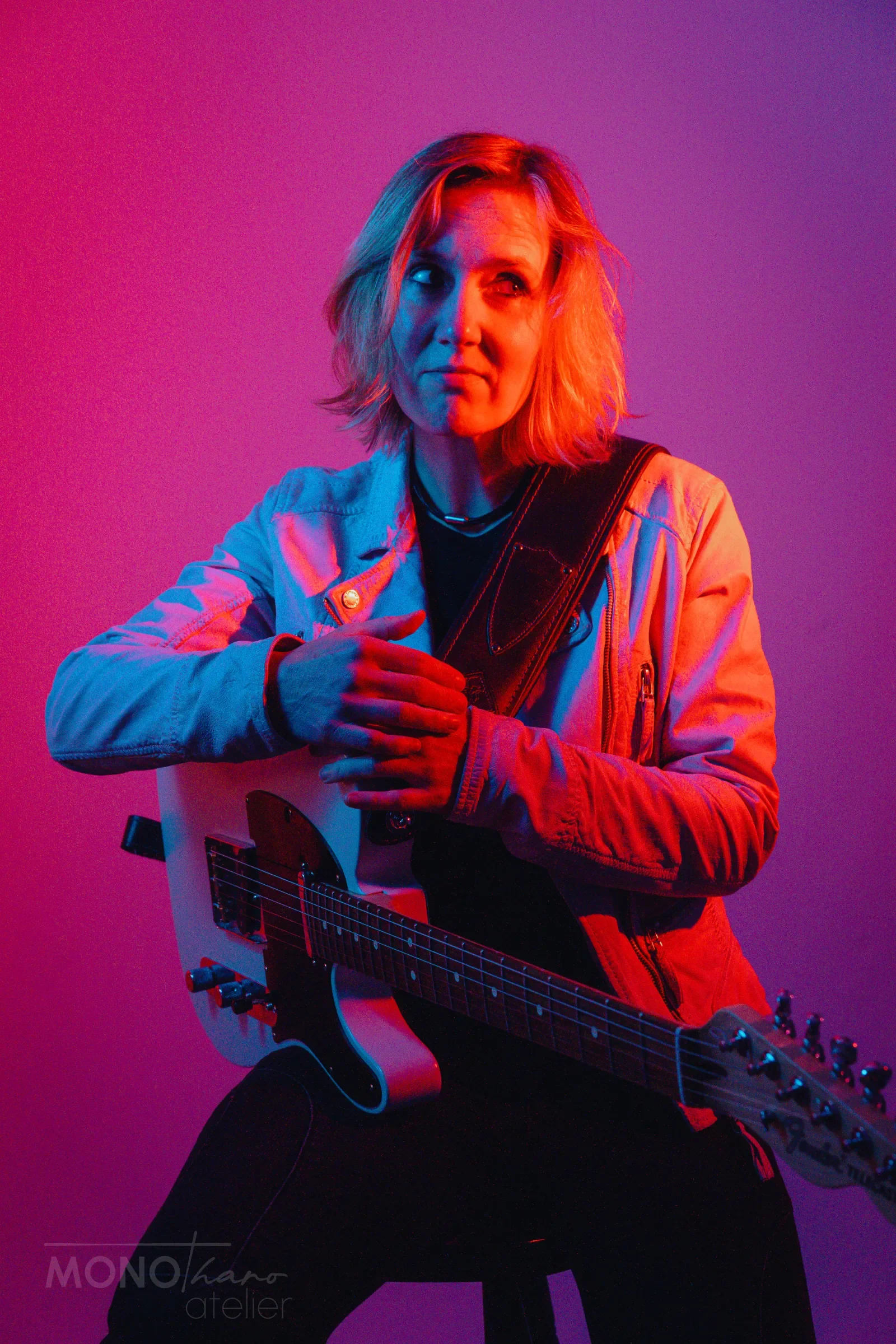 Eine Frau mit kurzen blonden Haaren spielt eine Akustikgitarre vor einem farbigen Lichthintergrund in Pink, Blau und Violett.