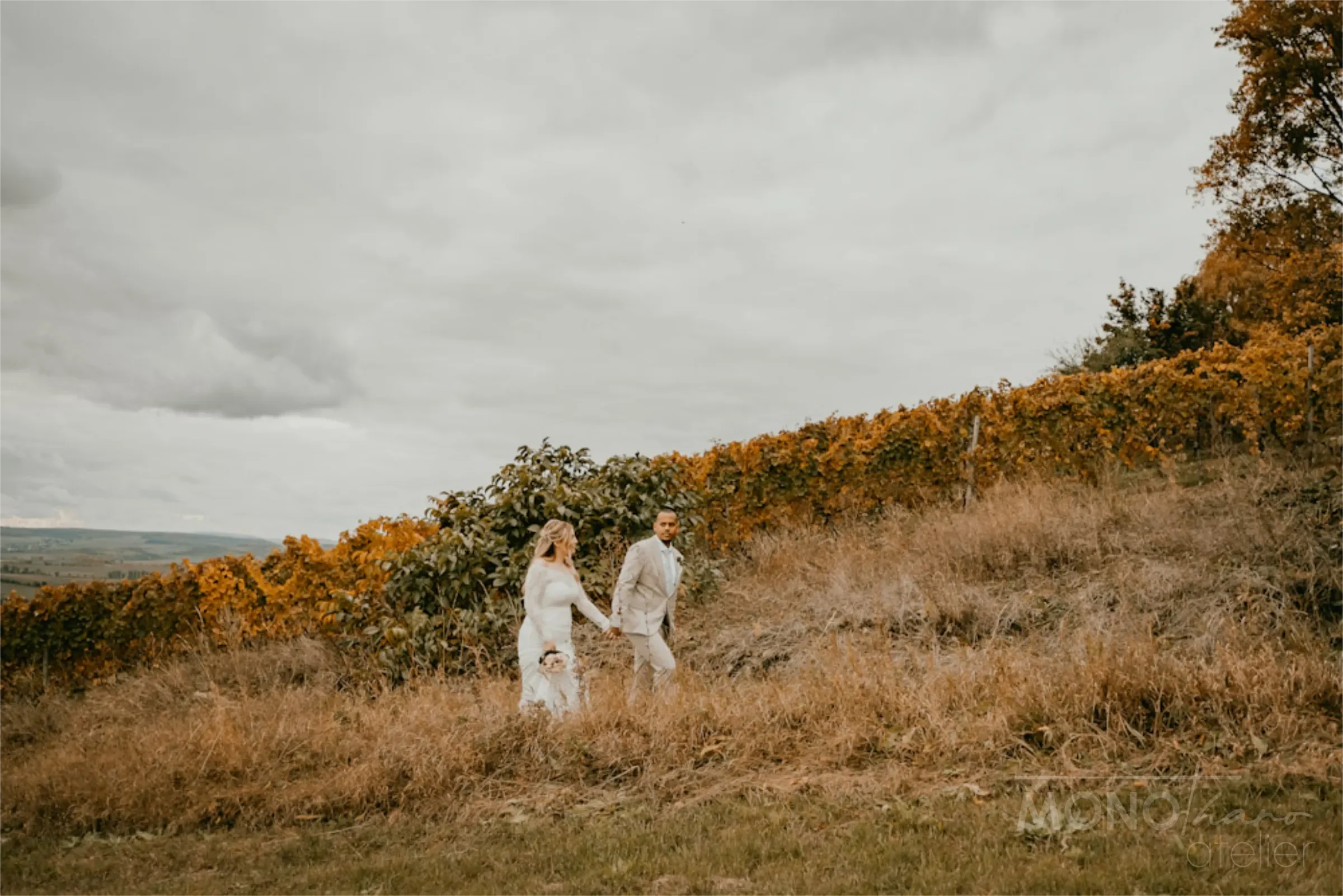 Ein Paar hält Händchen beim Wandern durch eine hügelige Landschaft mit Herbstbäumen und Wolken am Himmel.