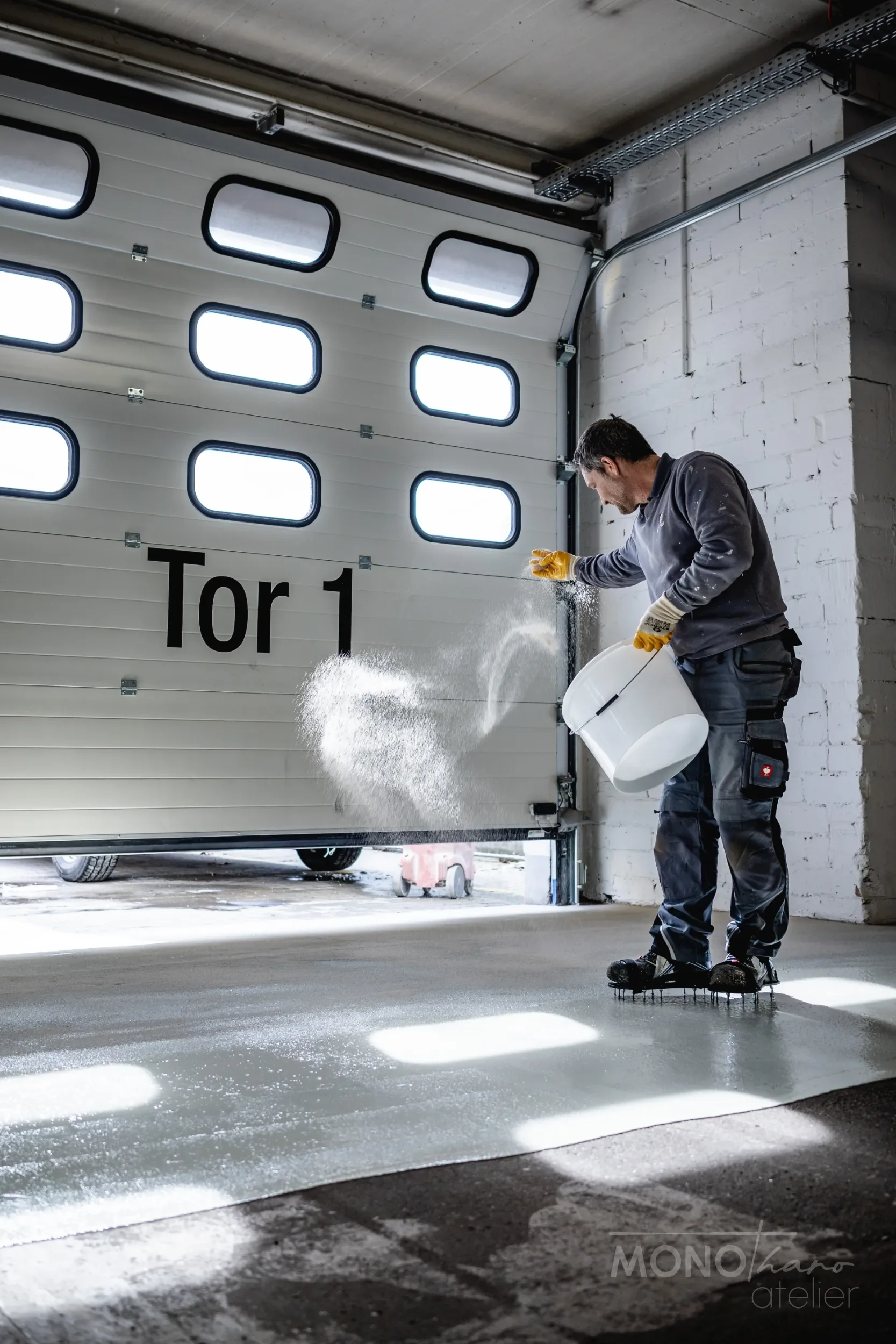 Ein Mann beim Schneeschaufeln in einer Garage, trägt schwarze Schuhe, eine graue Jacke und gelbe Handschuhe, und hat einen Eimer in der Hand.
