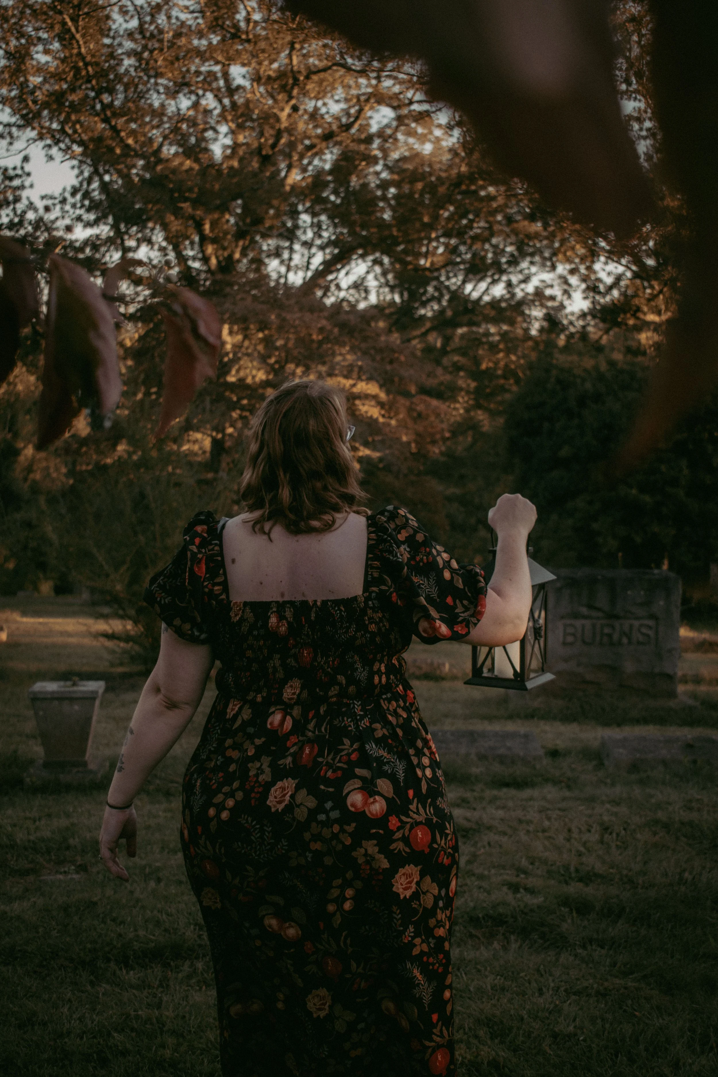 A woman with shoulder-length hair wearing a black floral dress with red and orange flowers walking in a cemetery at dusk. The woman is holding a lantern, and there are gravestones visible including one that has the word 'BURNS' on it. The background features tall trees with orange and brown leaves.