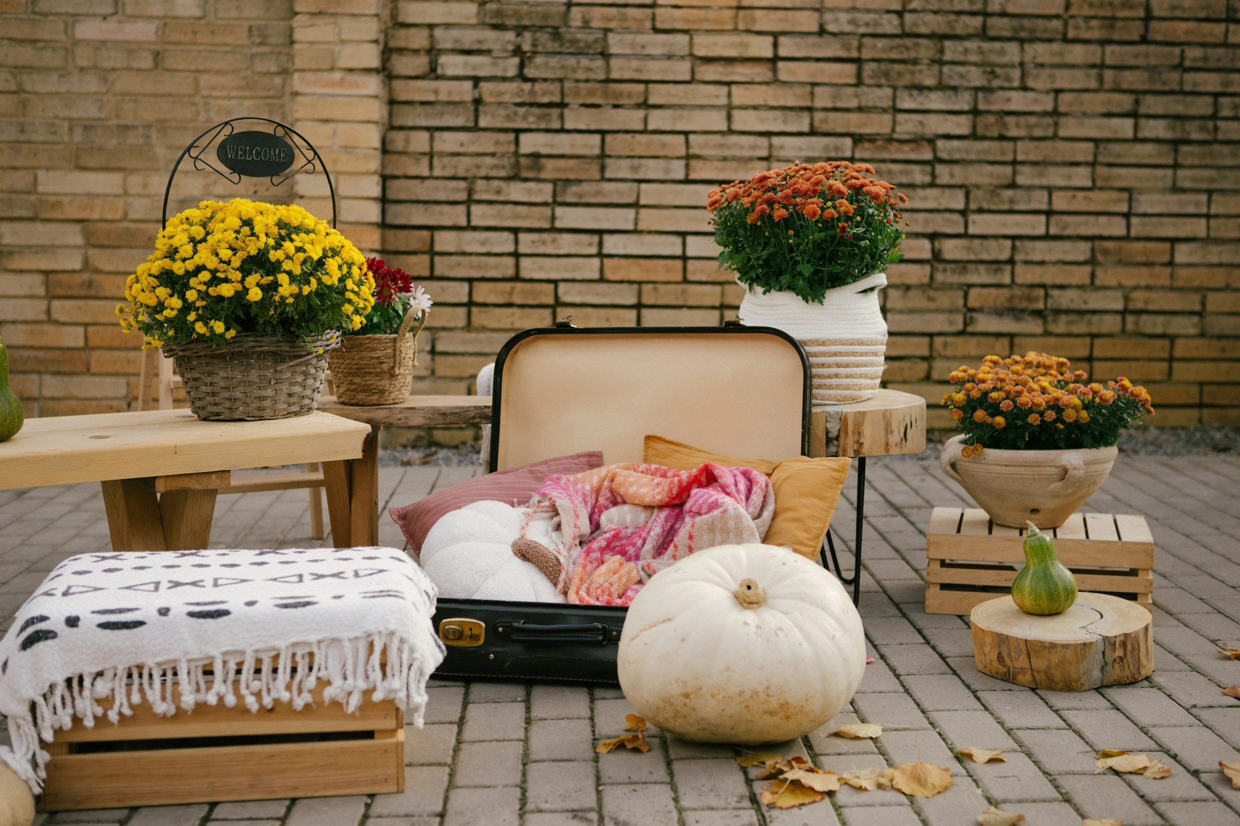 Outdoor scene with various planters of colorful flowers on a patio against a brick wall, with a woven basket and a wooden stool and pumpkins in the foreground.