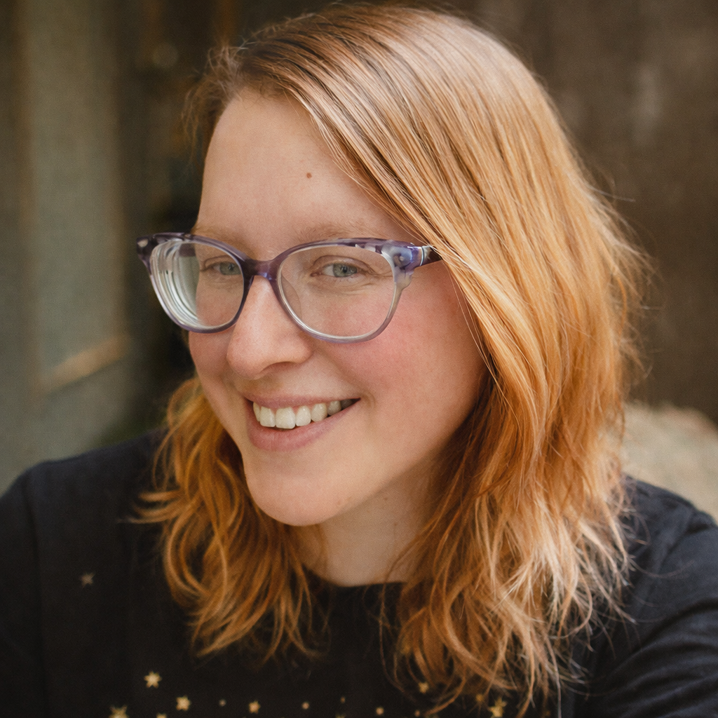 A young woman with red hair and glasses smiling outdoors.