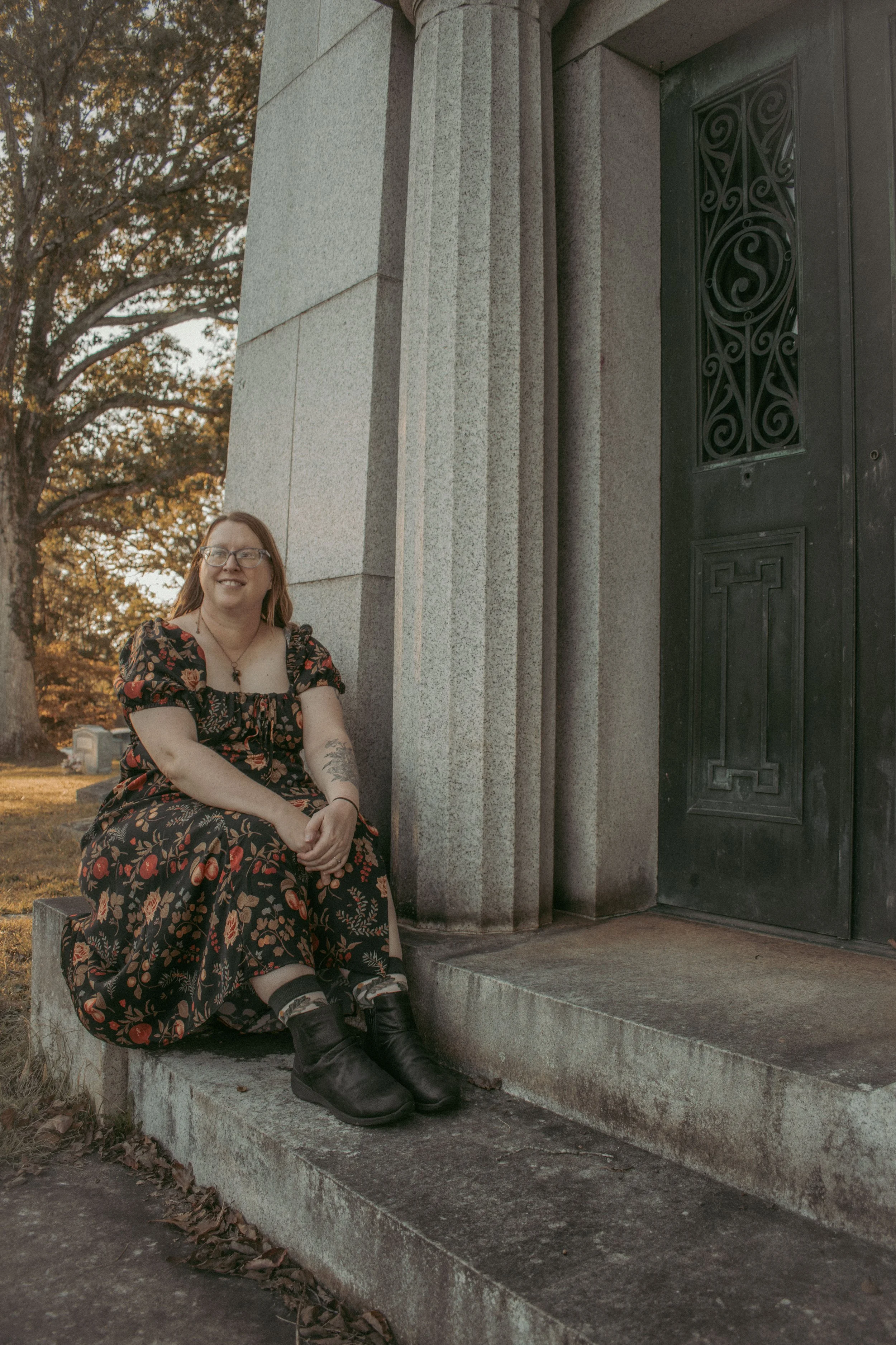 A young woman with glasses, wearing a black dress with red floral patterns, sitting on the steps of a stone building with a black ornate door, during autumn.