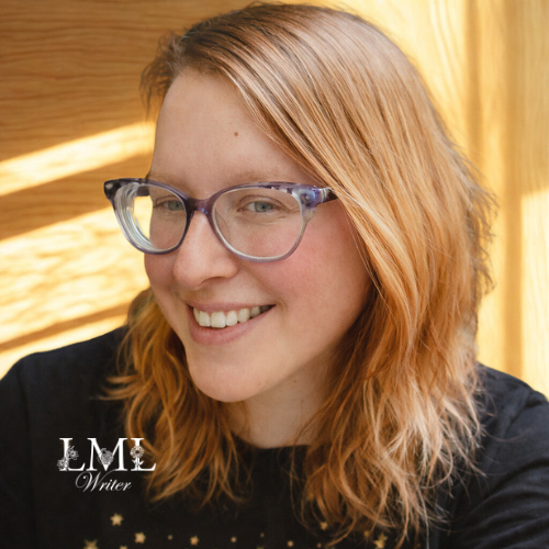 A woman with shoulder-length red hair and glasses smiling at the camera, wearing a black top, in a room with wooden and yellow curtains.