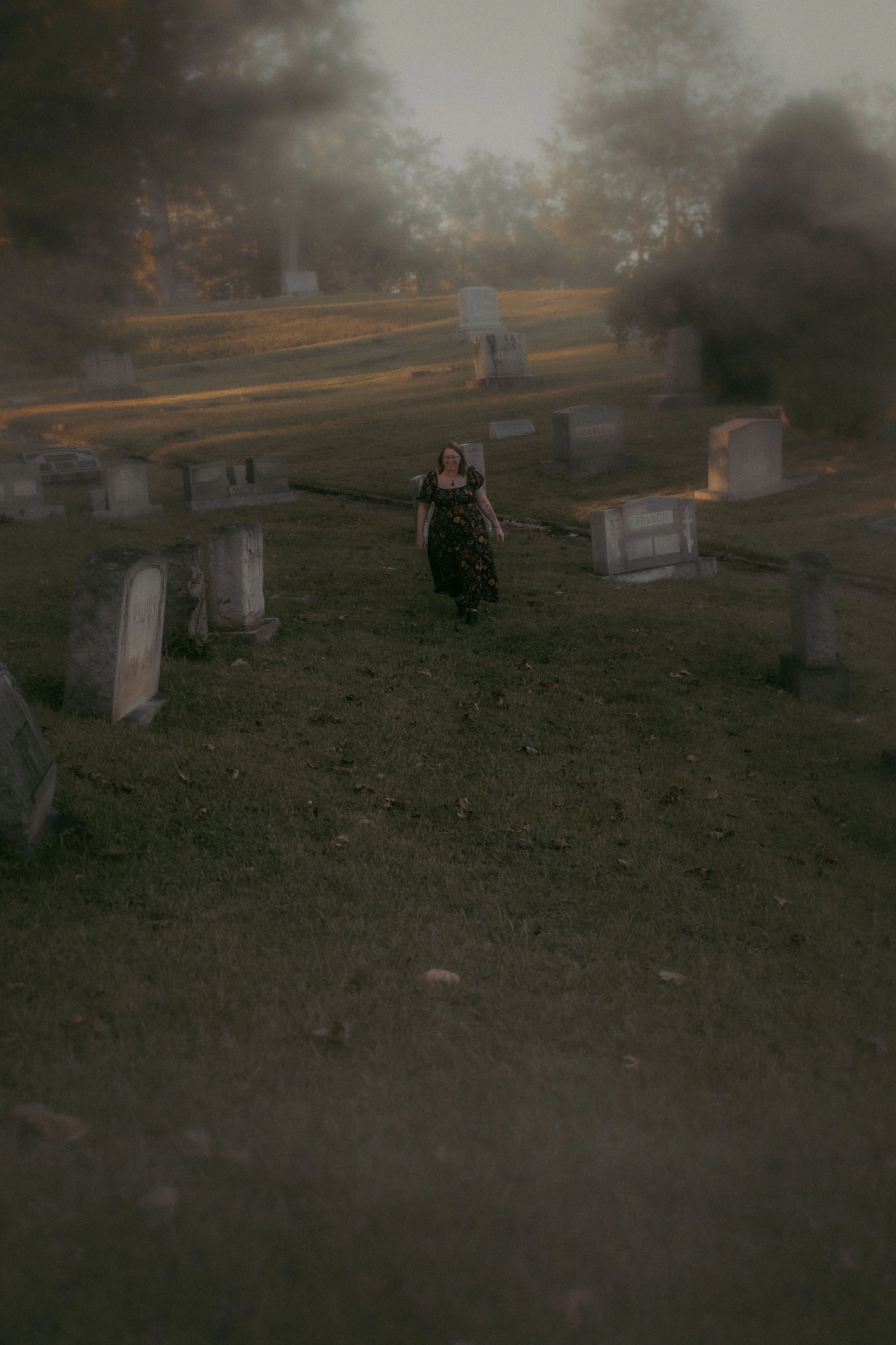 A woman walking through a graveyard with headstones during sunset, surrounded by trees and soft lighting.