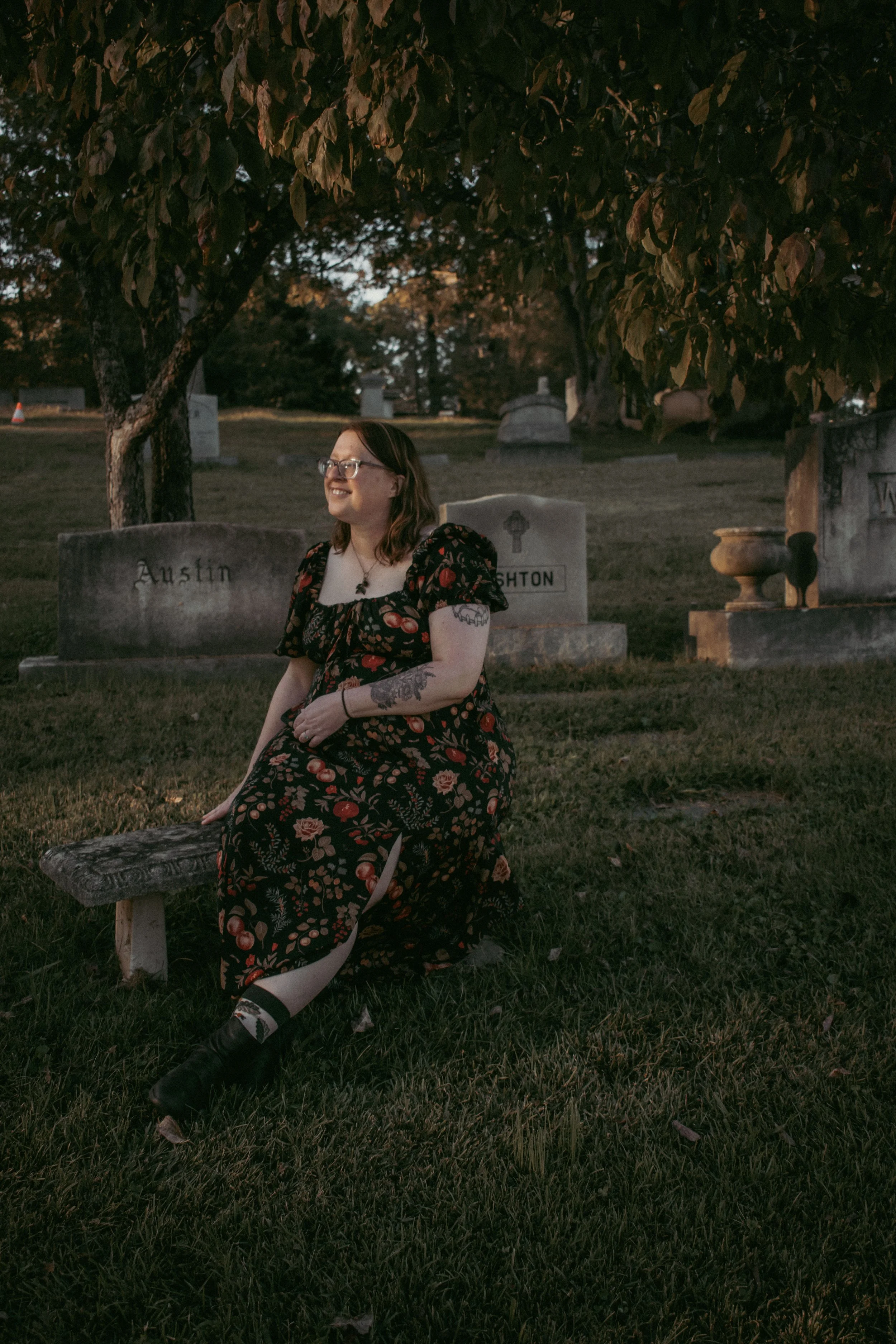 A woman with glasses and tattoos sitting on a park bench in a cemetery at dusk, wearing a black dress with a cherry pattern and smiling.