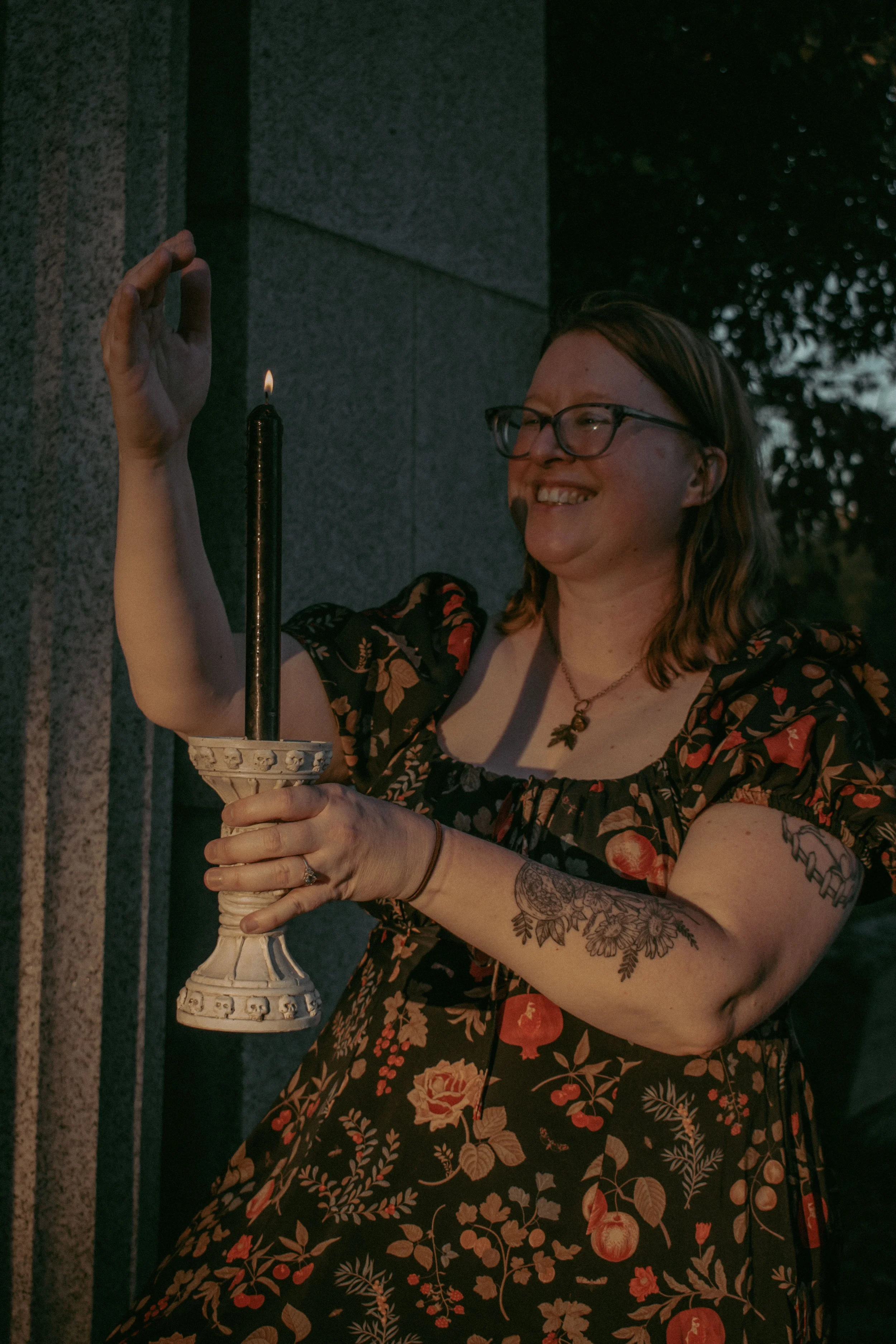 A woman with glasses, tattoos, and a floral dress holding a lit candle with a dark background.