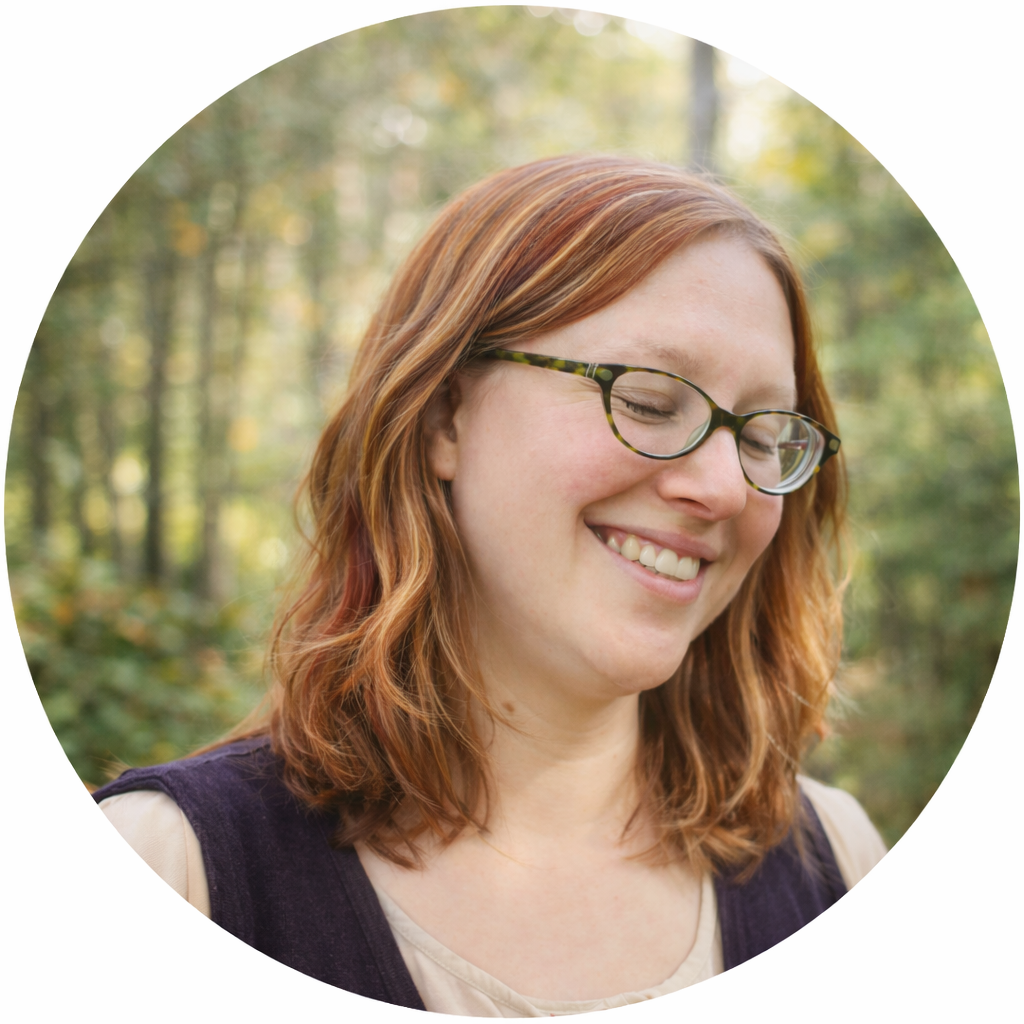 Smiling woman with glasses and red hair outdoors in a wooded area.
