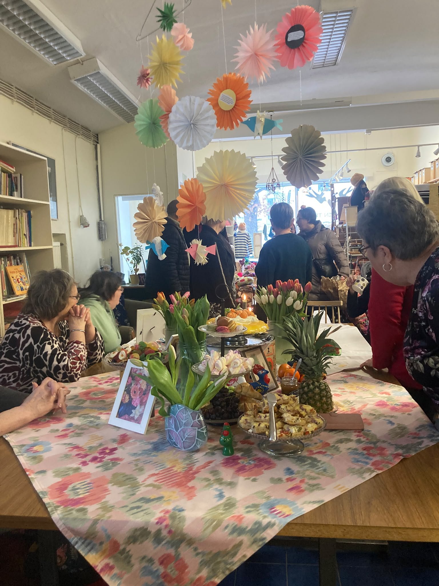 A lively indoor gathering with people around a table decorated with tulips, pineapples, and pastries. Colorful paper lanterns and flowers hang from the ceiling, creating a festive atmosphere.