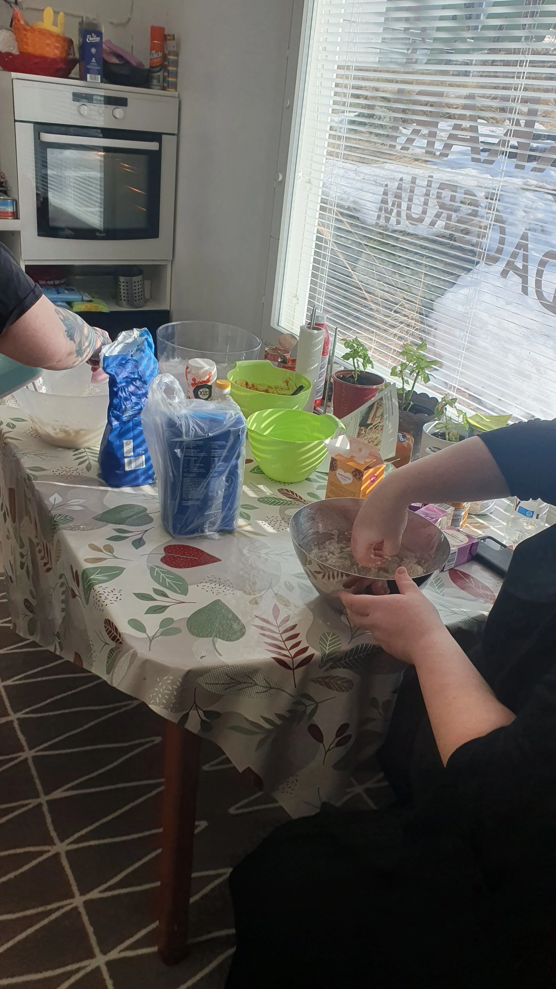 Henkilöt valmistavat ruokaa keittiössä, jossa on pöytä ja uuni. Pöydällä on erilaisia keittiötarvikkeita ja ainesosia.