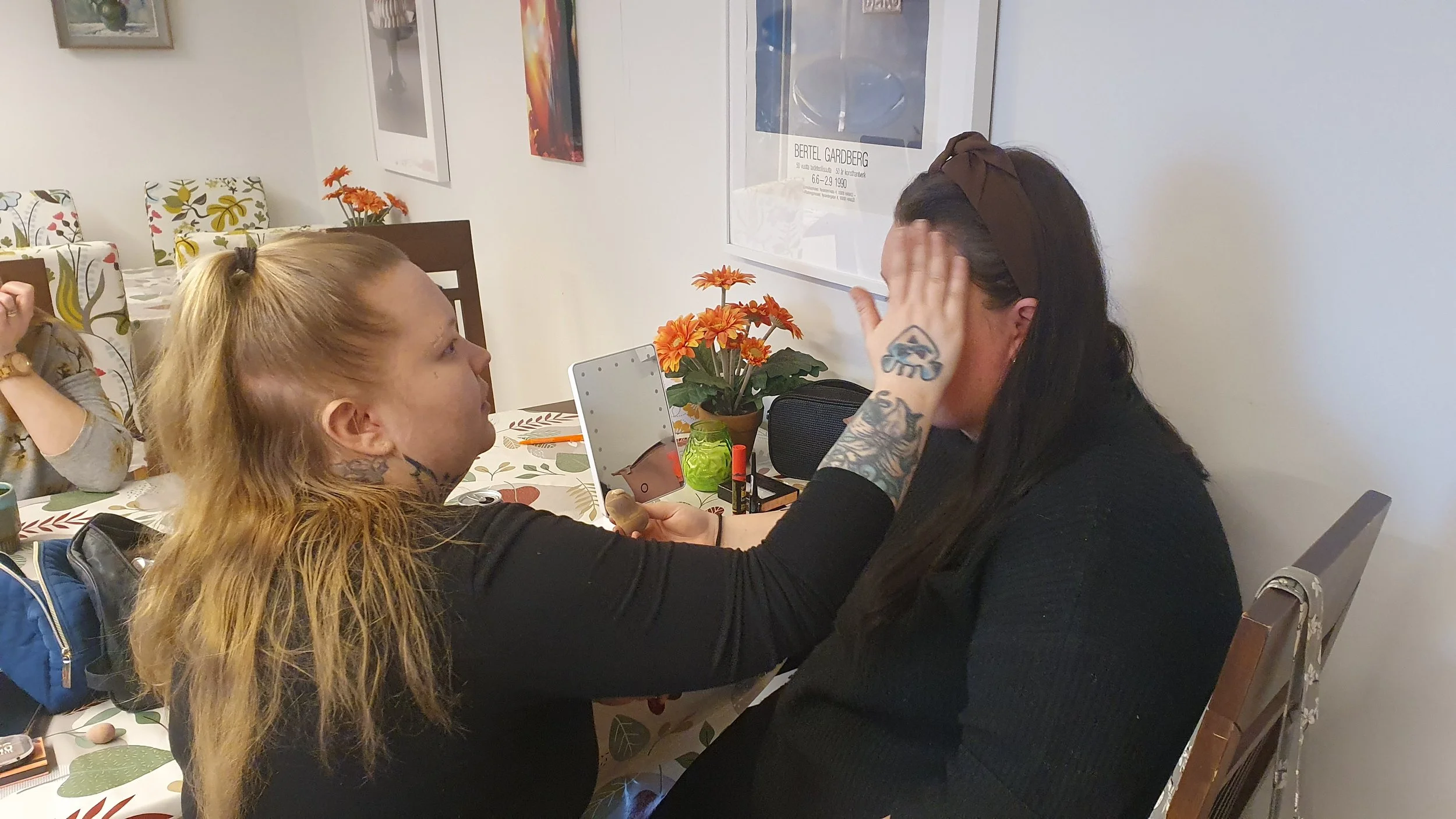 Kaksi naista on istumassa ruokapöydän ääressä, toinen tekee meikkiä toiselle.