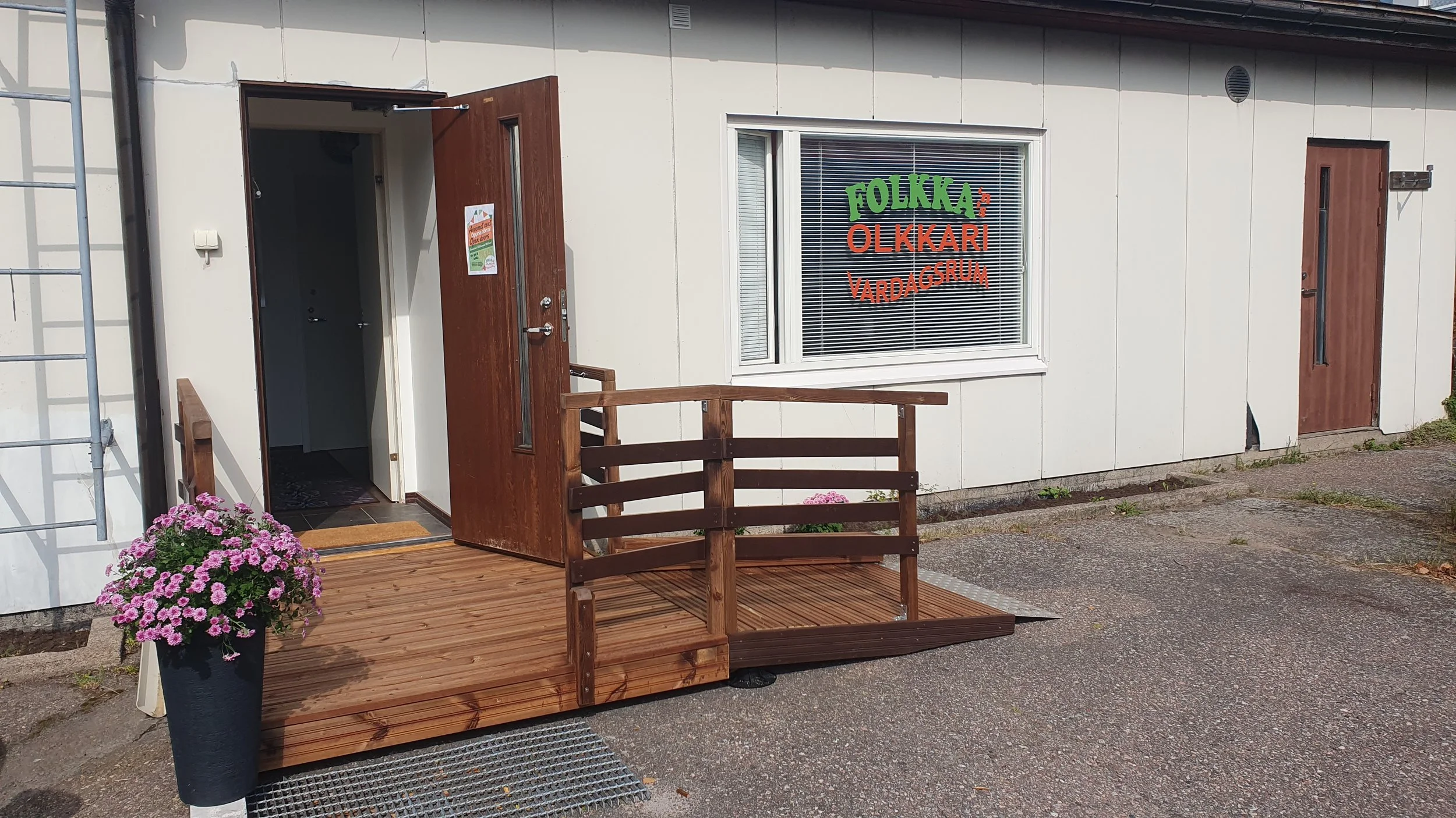 Kuvassa etualalla on pieni terassi, jossa on kukkaistutuksia ja puinen portaikko. Taustalla on rakennuksen ikkuna ja ovi, jonka ikkuna on koristeltu värillisillä kirjaimilla, jotka muodostavat tekstin 'FOLKKA OLLKARI VARDAGSFRUM'.