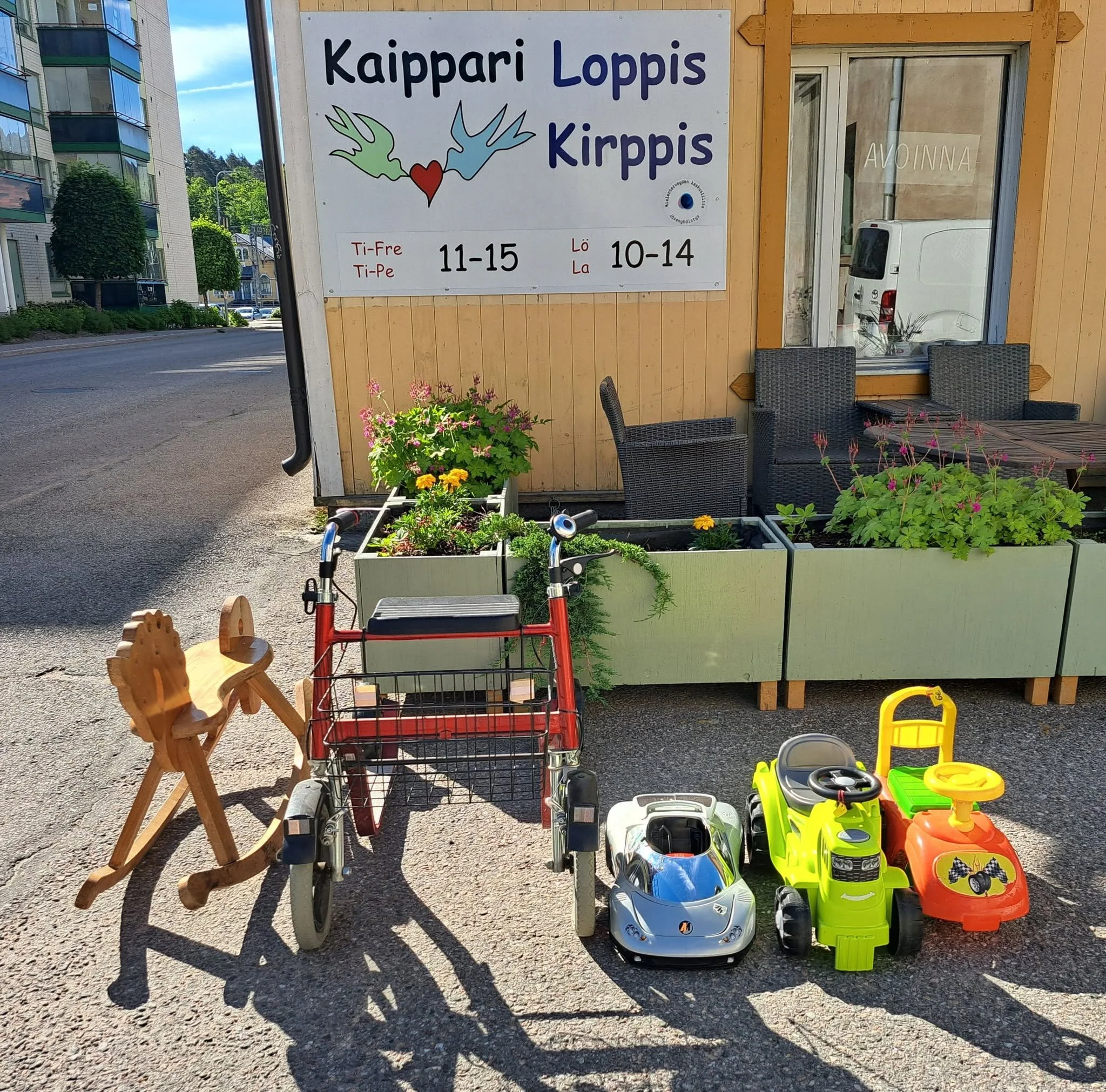 Kävellen myytävät lelut ja lastenajoneuvot ulkona. Taustalla on vaalea puutalo ja ostoskeskus.