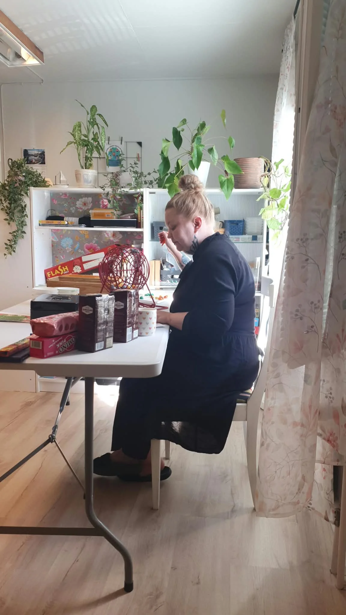 Nainen istuu ruokapöydän ääressä keittiössä, ympärillä joululahjaesineitä ja kakkuja, taustalla kasveja ja kaappi.