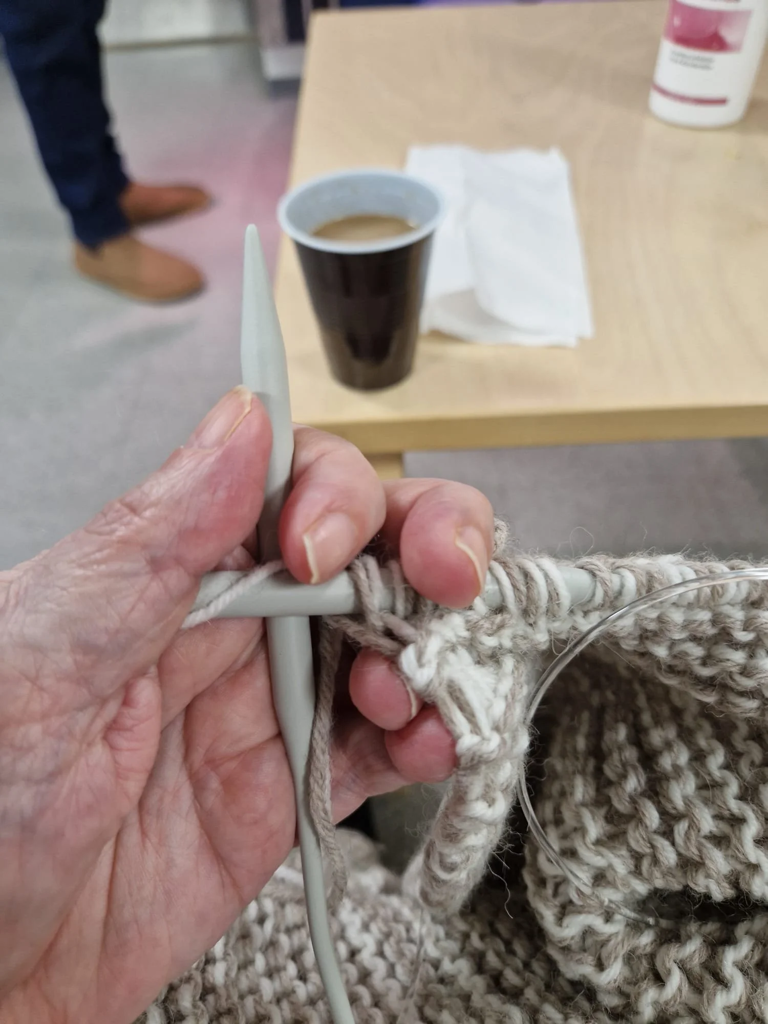Kuvassa ihmisen käsi, joka pitää piirtopäistä ja istuu pöydän ääressä. Pöydällä on muovimuki, joka sisältää juomaa, ja paperinen lautasliina. Taustalla näkyy muovipullo ja toisen ihmisen jalat, jotka on pukeutunut farkkuihin ja vaaleanruskeisiin kenkiin.