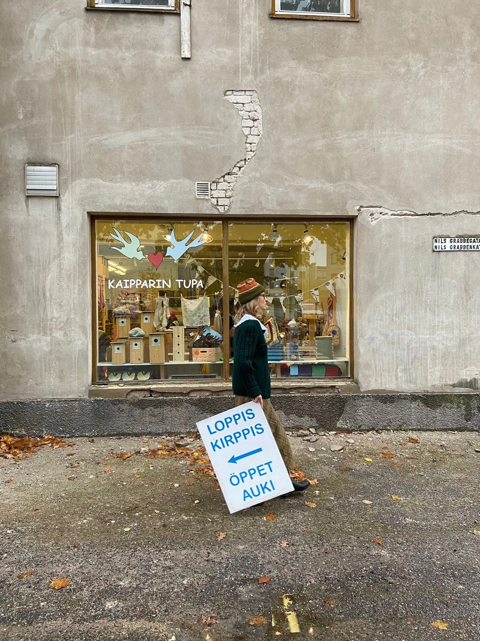 Keskellä nainen, joka kävelee poispäin, kantaa valkoista lappua, jossa lukee 'LOPPIS KIRPPIS'. Taustalla on ikkunaseinä, jossa on kauppaliikkeen nimi 'KAIPPARIN TUPA' ja myynnissä olevia tavaroita näkyvissä.