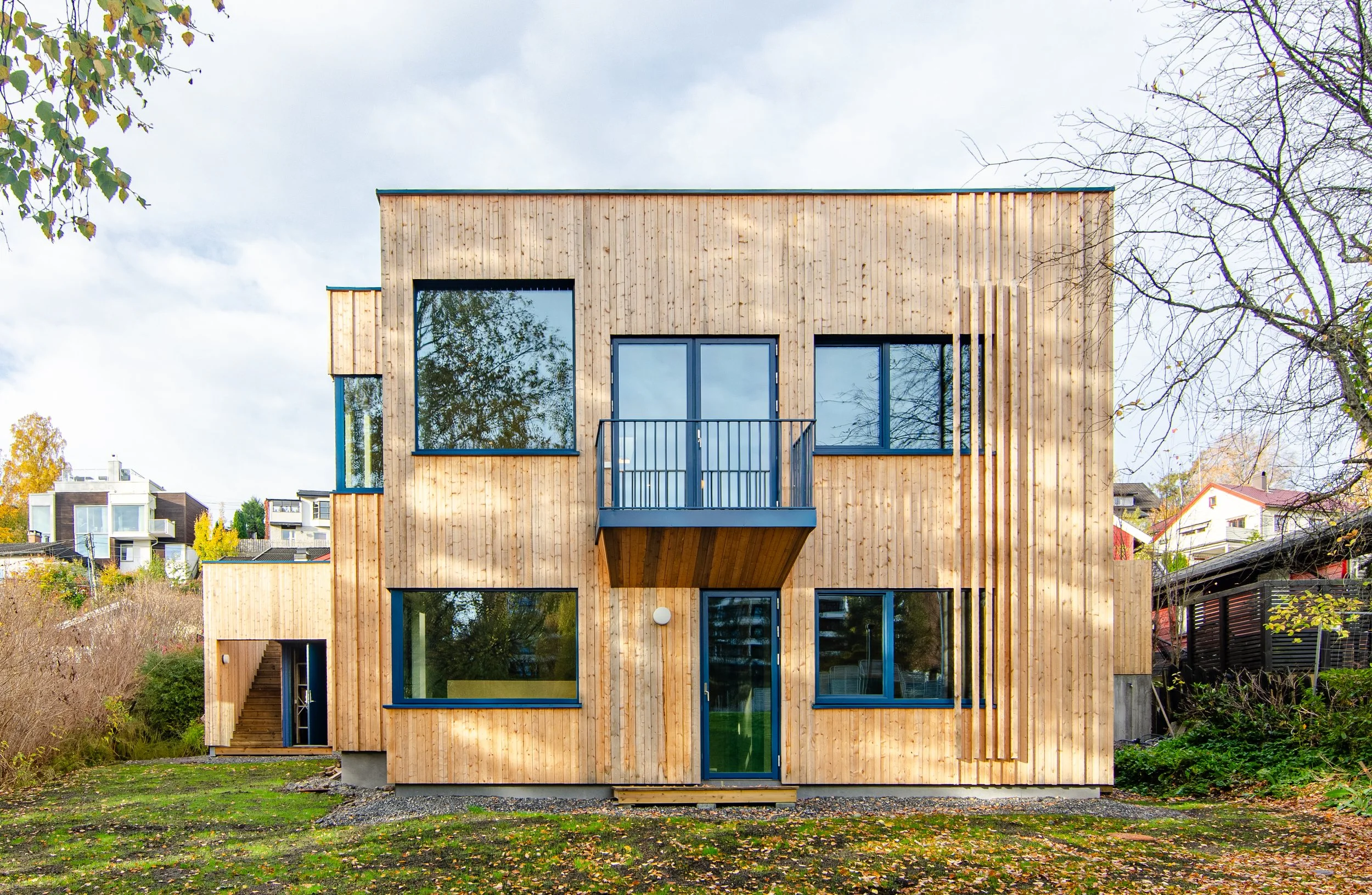 Modern wooden multi-story house with large windows, a small balcony, and surrounded by trees and greenery.