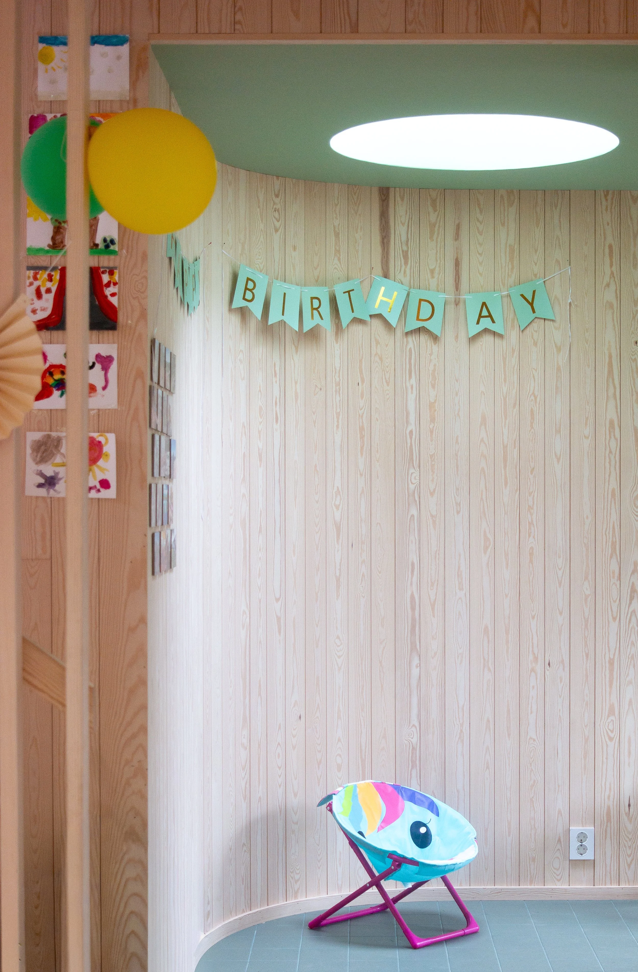 A wooden-paneled indoor space decorated for a birthday party, with a green and yellow balloons, children's artwork on the wall, a light blue banner with the word 'BIRTHDAY,' and a colorful fish-shaped chair on the tiled floor.