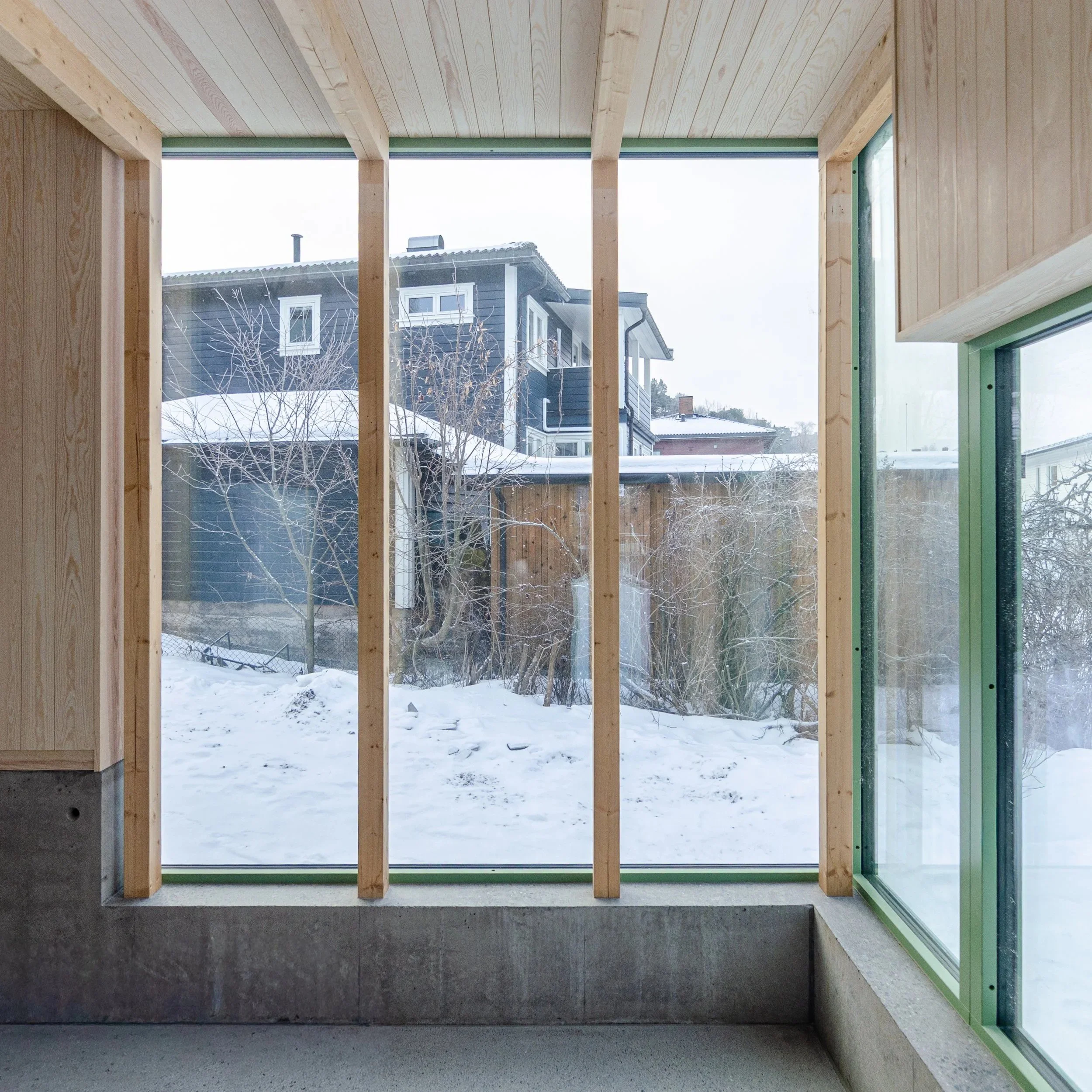Interior view of a partly constructed room with wooden framing and large windows overlooking a snowy residential neighborhood.