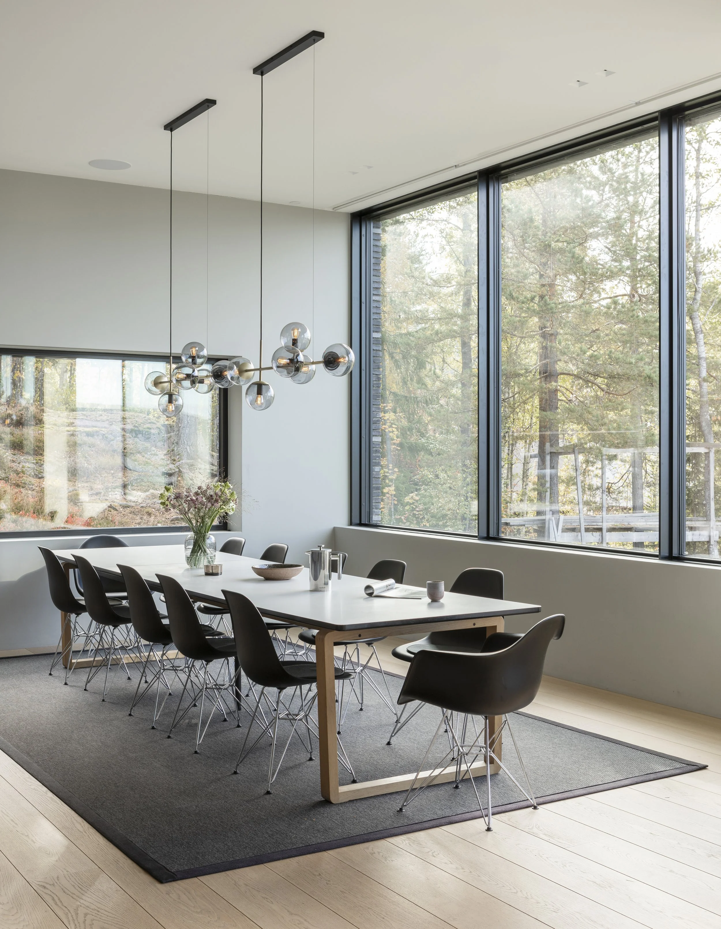 Modern dining room with large windows showcasing a forest view, black chairs around a white table, and a contemporary chandelier hanging from the ceiling.