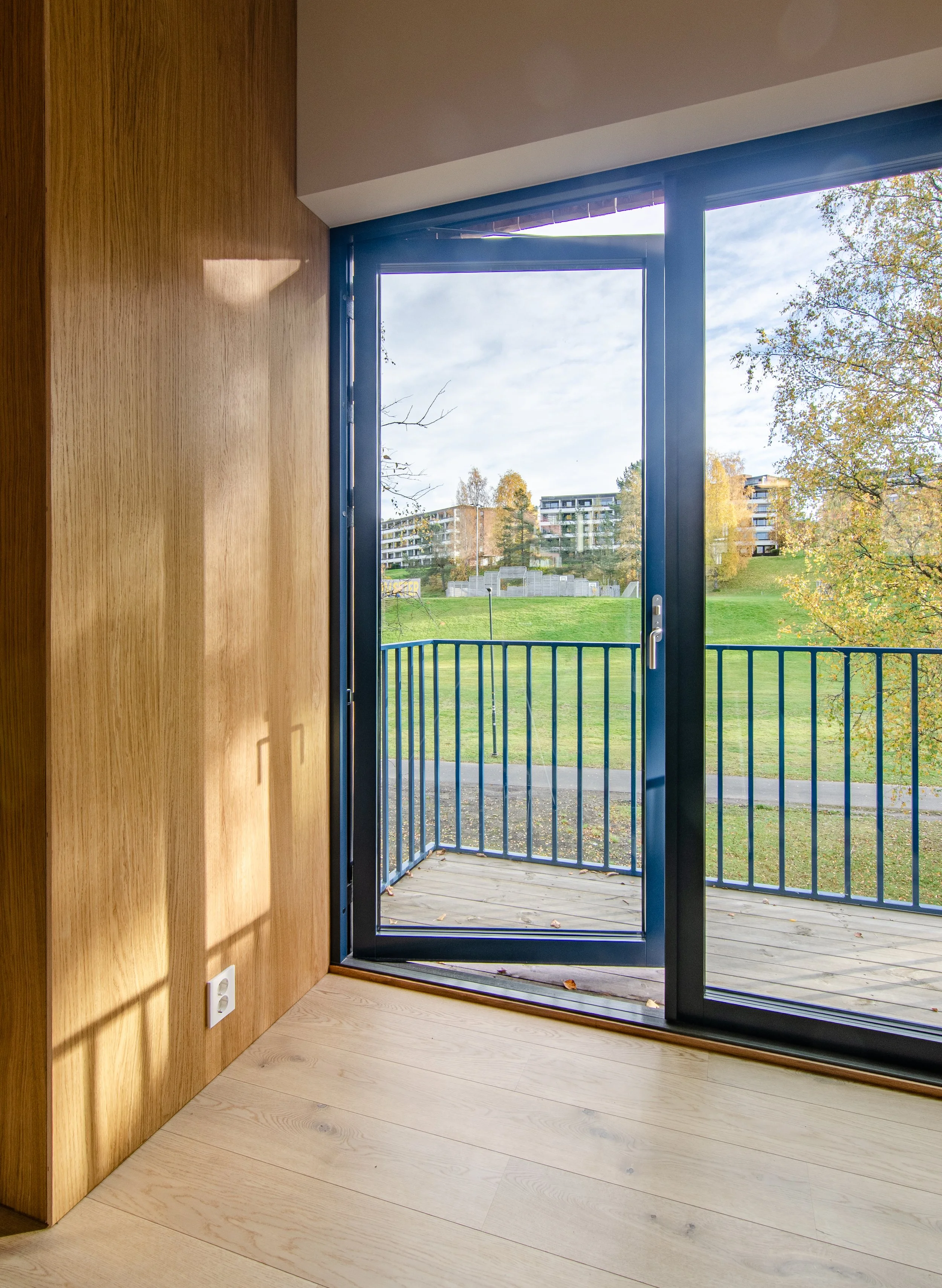 Interior view of a room with wooden wall paneling and a sliding glass door opening to a balcony overlooking a grassy area with trees and apartment buildings in the background.
