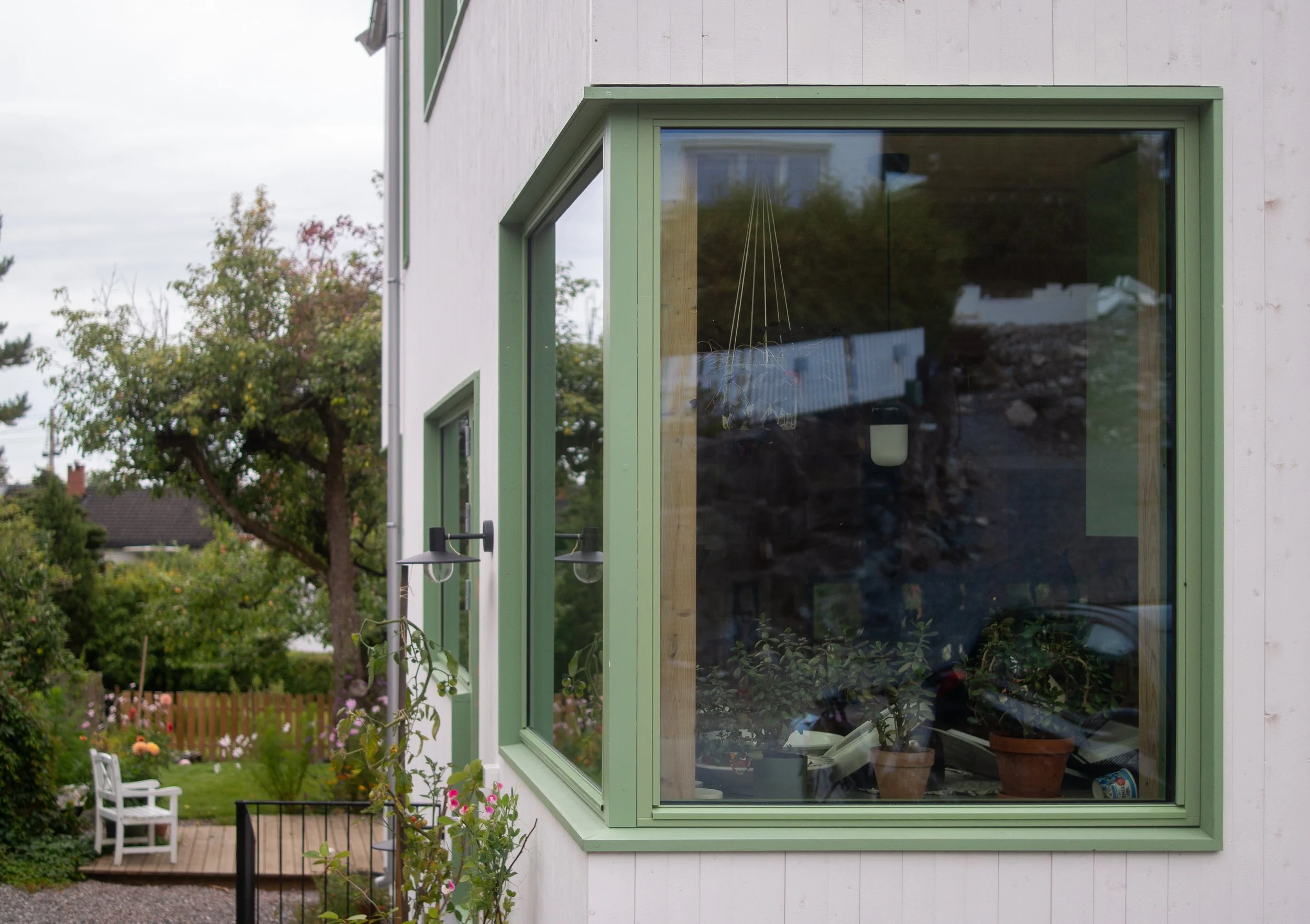 Front corner of a house with a large glass window and green trim, showing indoor plants inside. Outside, there is a garden with flowers, a white chair, and trees in the background under an overcast sky.