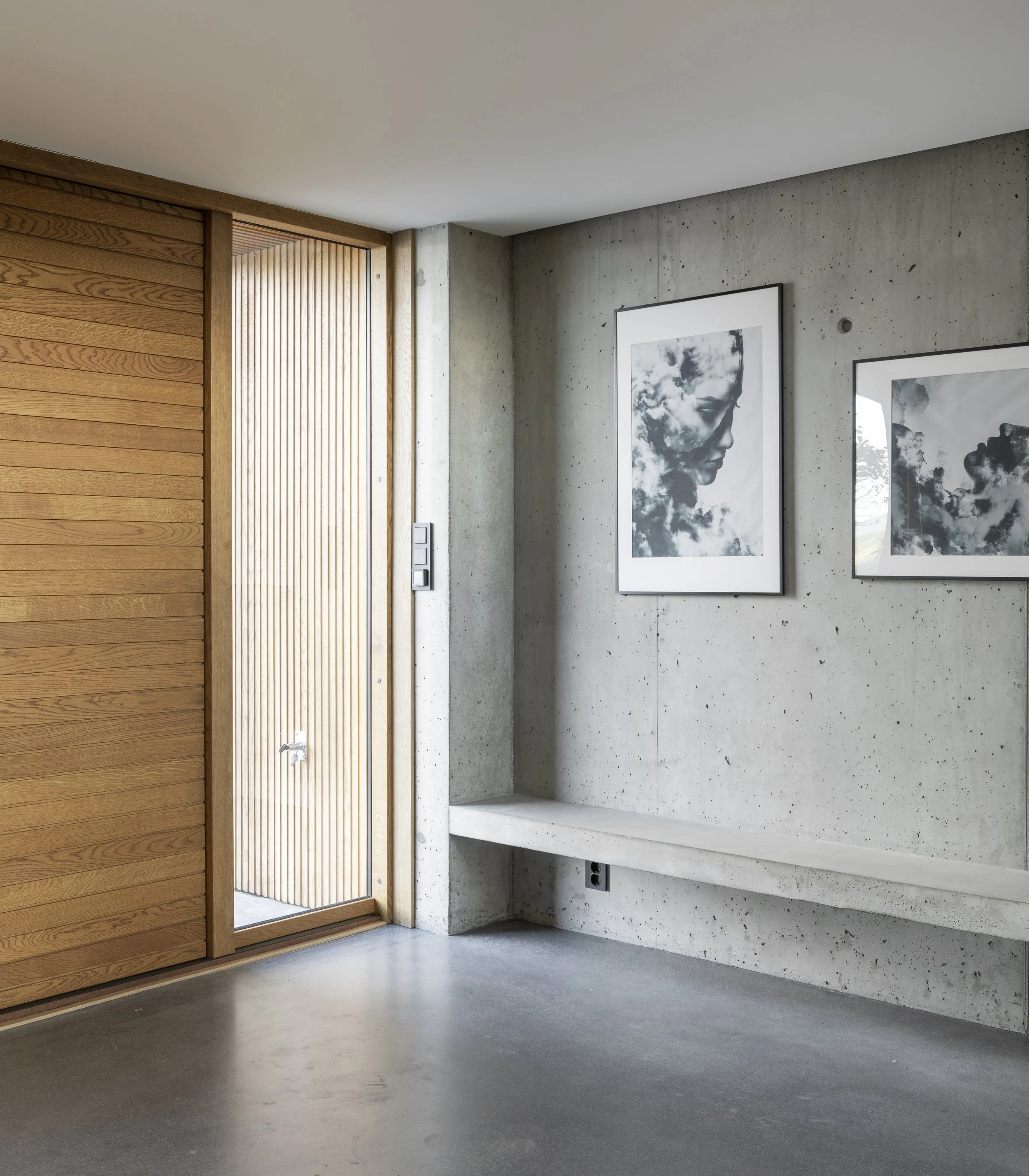 Modern interior with wooden slats, concrete walls, two framed black-and-white portraits, a large window, and a polished concrete floor.