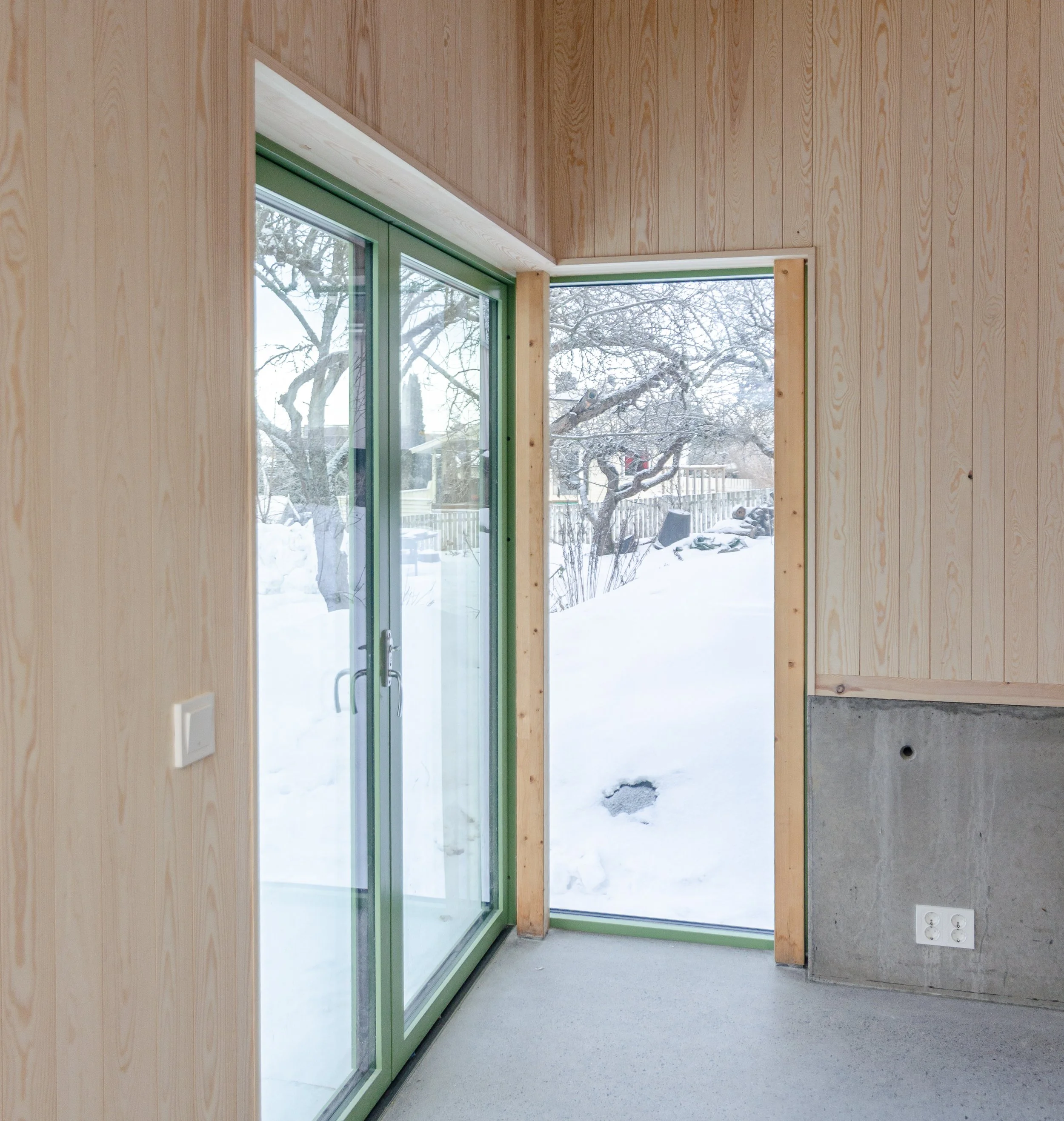 Interior of a room with unfinished wood walls, glass sliding door with green frame, view of snow outside, and electrical outlets on the wall.