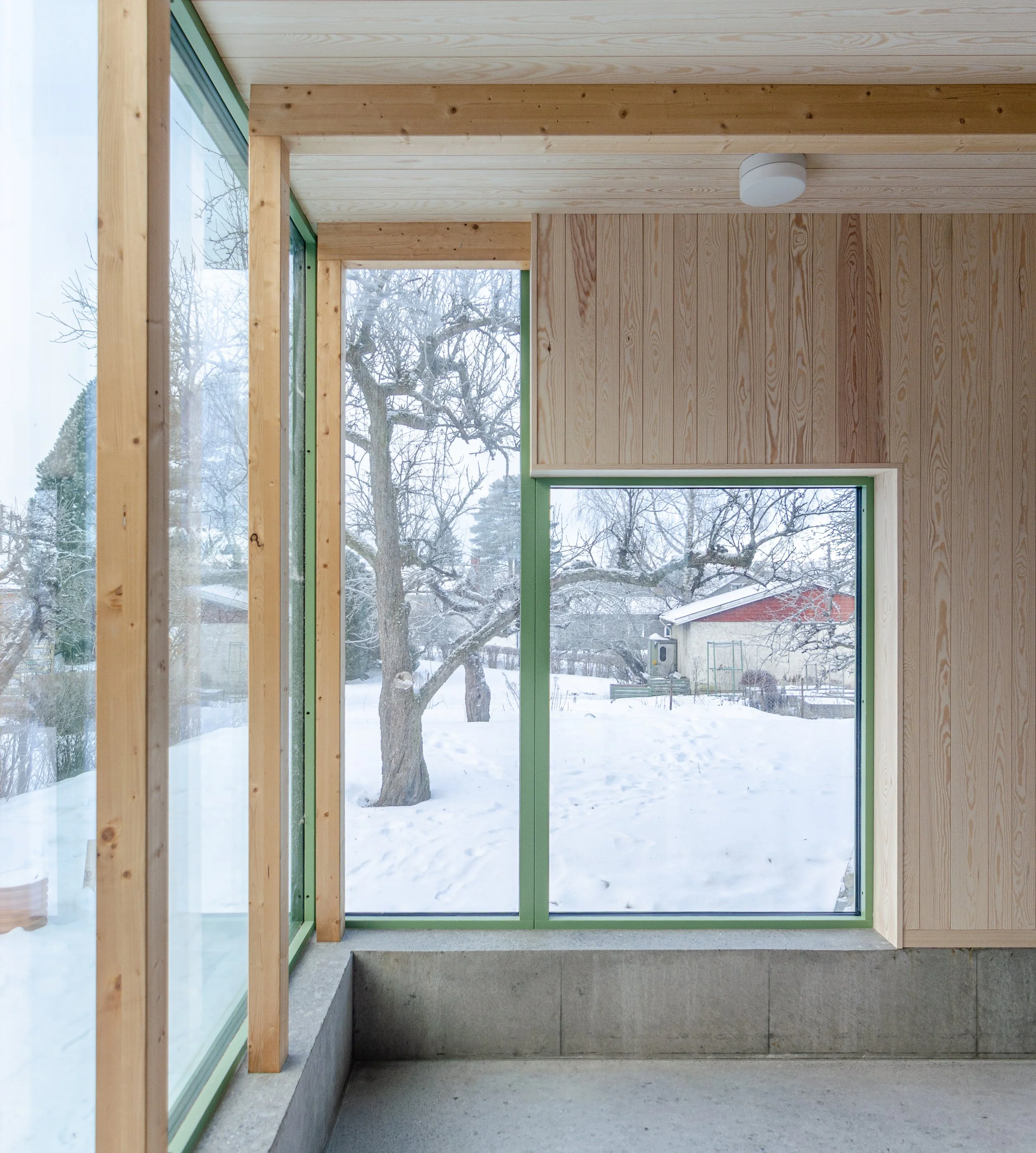 Interior view of a room under construction with large windows showing a snowy outdoor landscape, including trees and a red building. The room has wooden wall paneling, a green window frame, and an unfinished concrete floor.
