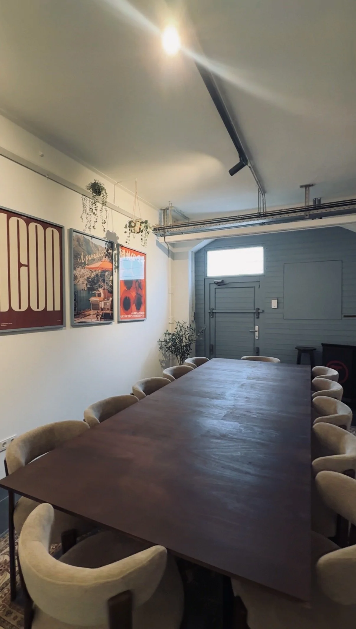Ein Konferenzraum mit einem langen Holztisch, umgeben von beige, bequemen Stühlen. Die Wände sind mit Postern und Pflanzen dekoriert, im Hintergrund eine Tür und ein Fenster. Das Zimmer hat ein modernes, gemütliches Ambiente.