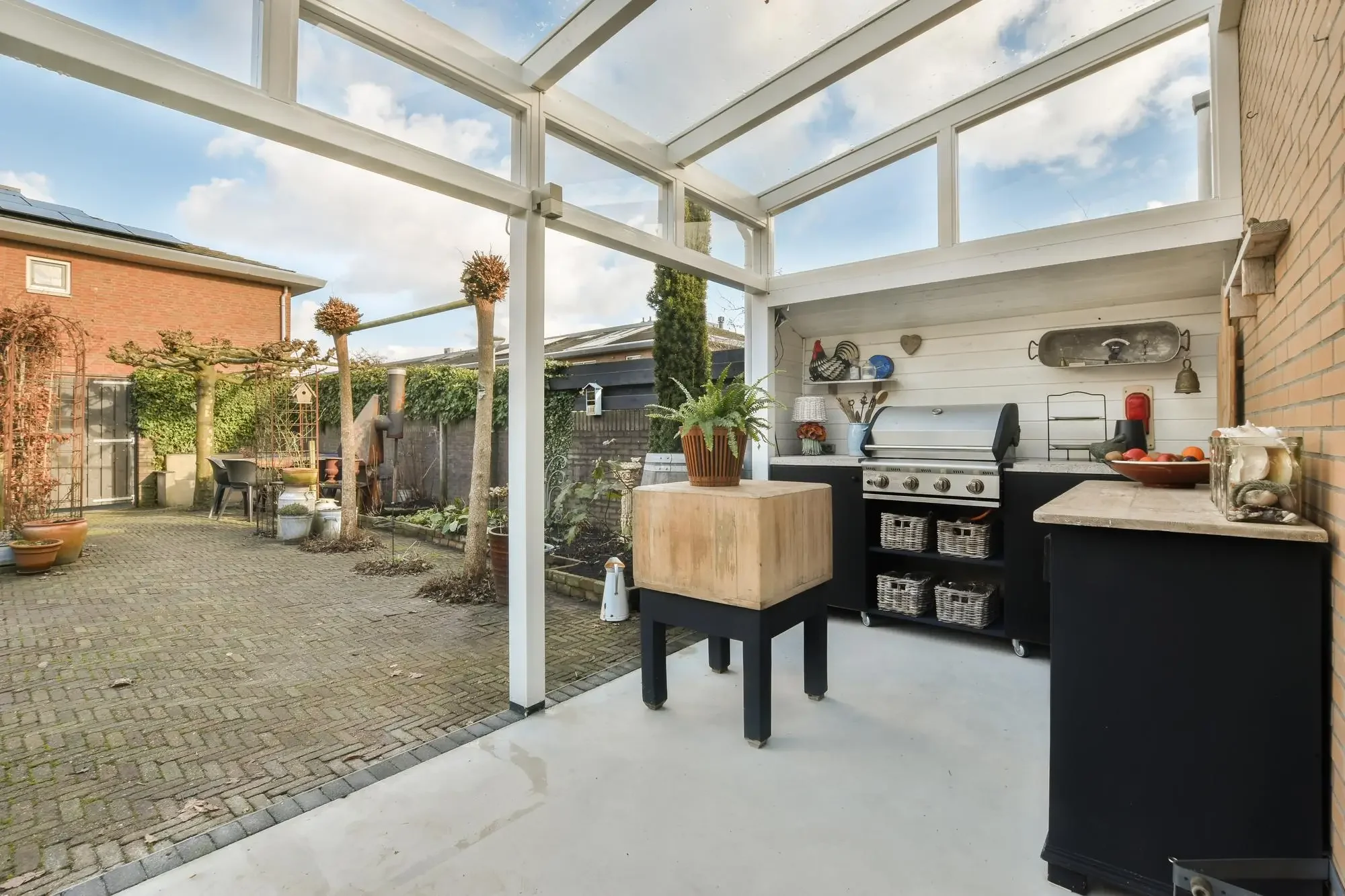 A backyard patio area with a covered outdoor kitchen and a garden. The patio has a modern black outdoor grill, a small wooden table, and potted plants. The garden includes trees, shrubs, and a brick fence. The sky is partly cloudy.