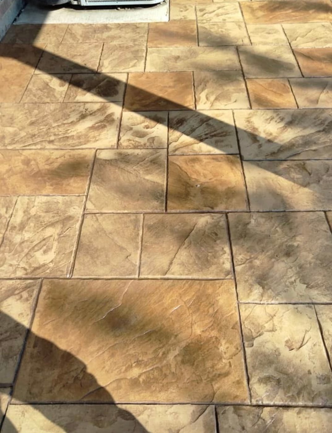 Close-up of a tiled outdoor floor with beige and brown stone-patterned tiles, patches of sunlight, and shadows on the surface.