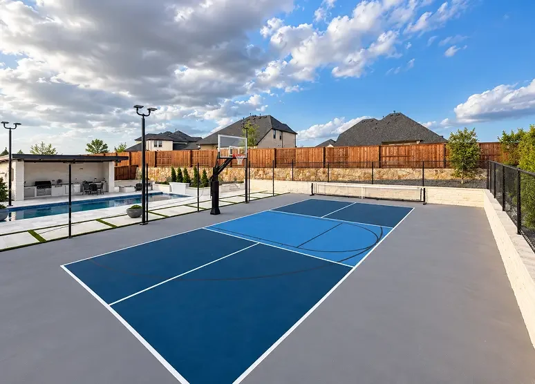 Outdoor basketball court with blue playing surface and white lines, next to a pool area with seating, surrounded by a wooden fence.