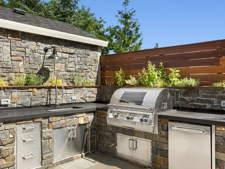 Outdoor kitchen with stone and brick finish, built-in grill, stainless steel cabinets, black countertop, garden bed with green plants, wooden privacy fence, and trees in the background.