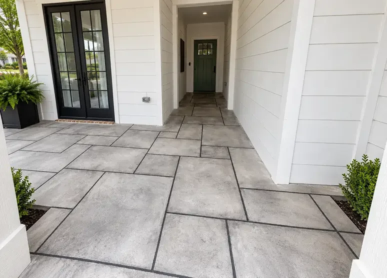 Covered porch with gray concrete tiles, black door with glass panes, and white siding, with green plants on either side.