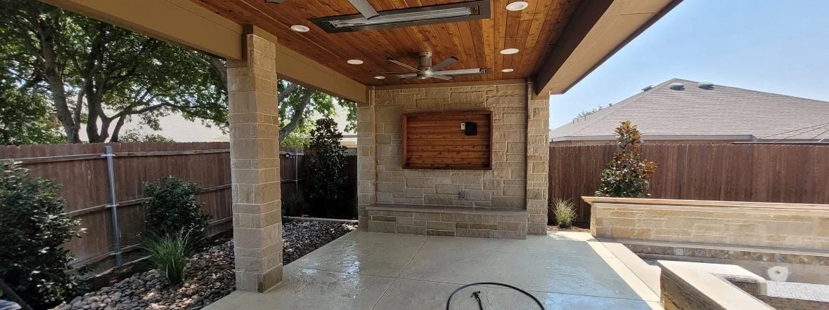 Covered outdoor patio with a wooden ceiling, ceiling fan, stone pillars, and a wooden wall mounting with a grill or TV mount. Surrounding a backyard with a wooden fence, small trees, and plants.