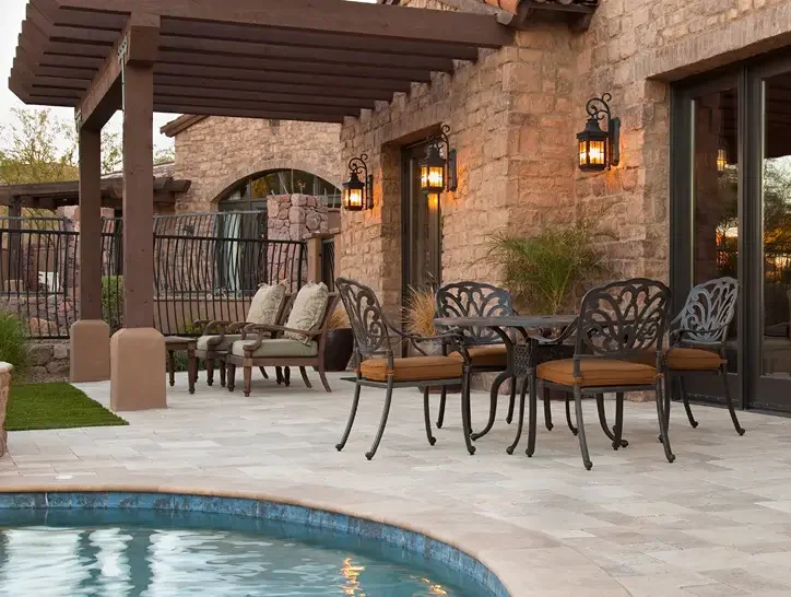 Outdoor patio area next to a swimming pool, with wrought iron chairs and tables, cushioned lounge chairs, lantern-style wall lights, and a brick building with large windows and a pergola.