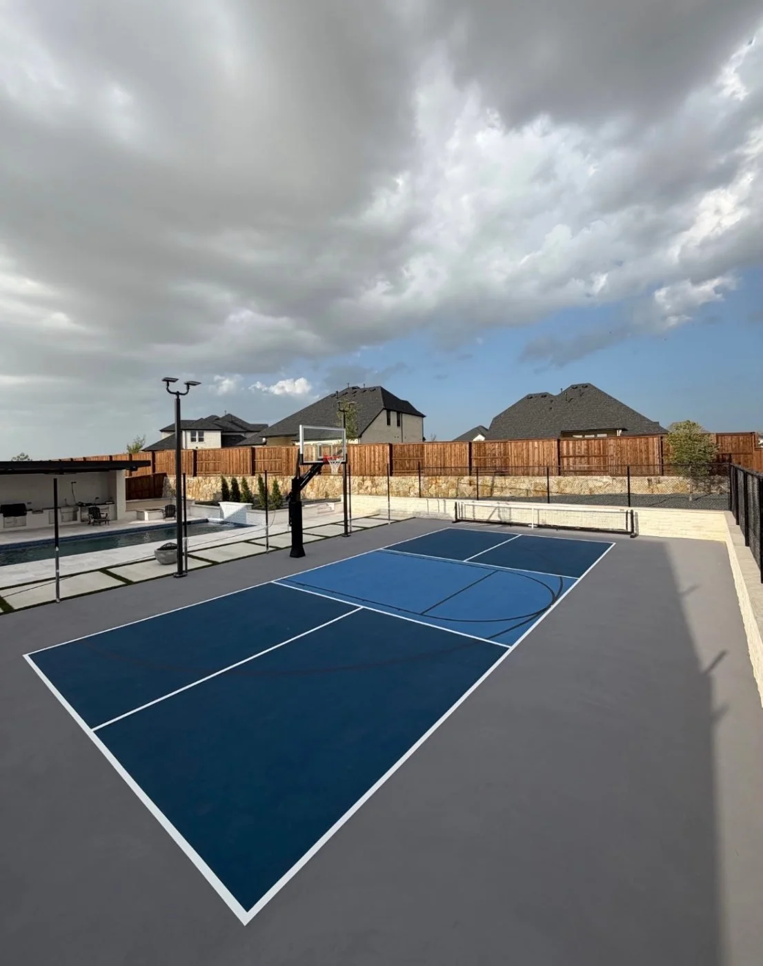 A modern outdoor basketball court with blue surface, black net, and fencing, along with a backyard pool and lounge area, under a cloudy sky.