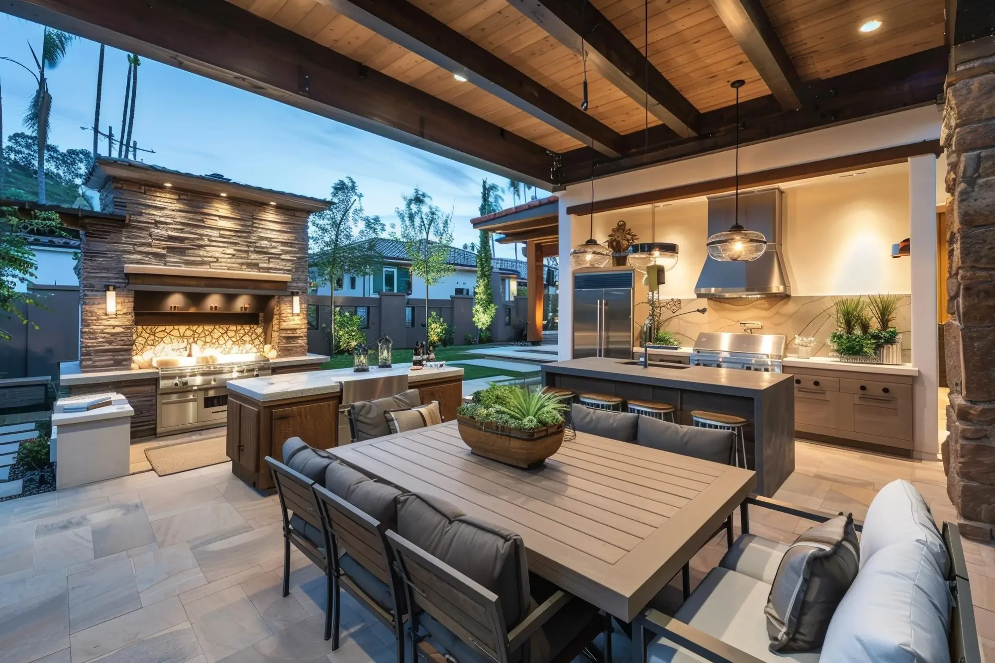 Outdoor living area with a dining table, chairs, and a kitchen with a grill under a wooden ceiling, with a backyard, trees, and neighboring houses visible.