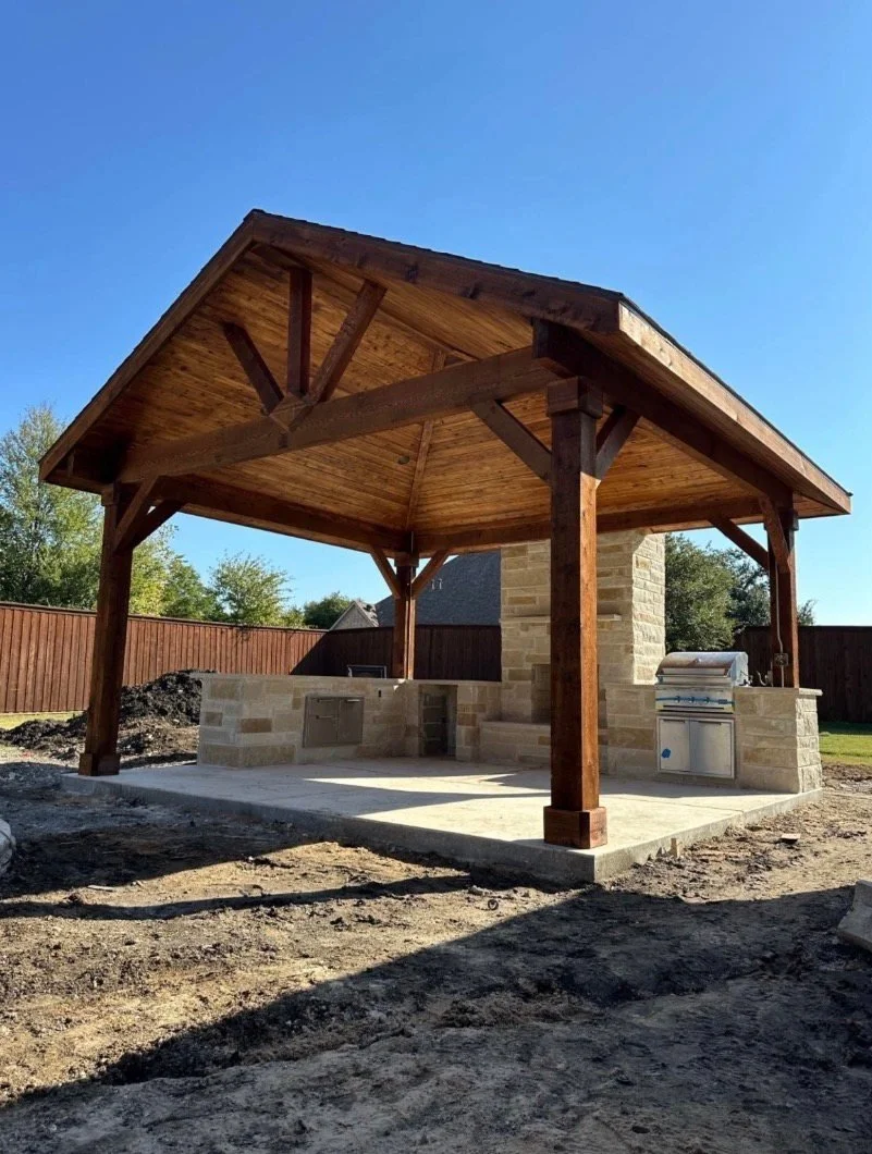 Outdoor covered kitchen with a stone base, built-in grill, and wooden roof and support beams, in a backyard with dirt ground and a wooden fence.
