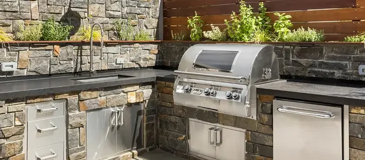 Outdoor kitchen with built-in stainless steel grill, stone countertops, and storage cabinets, surrounded by potted plants.