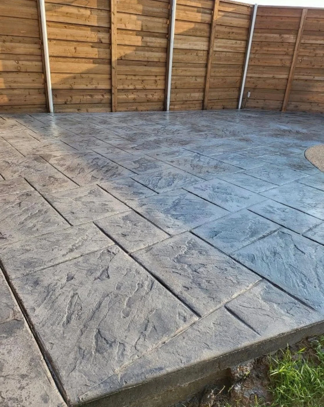 Newly installed gray stone-patterned concrete patio in a backyard with a wooden privacy fence.