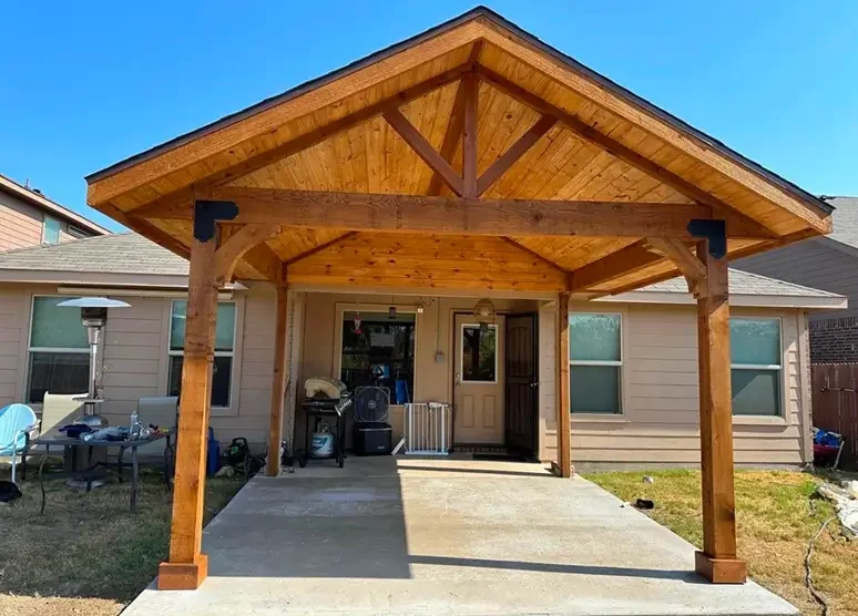 Back patio of a house featuring a newly constructed wooden pergola with a gabled roof, supported by four sturdy wooden posts, standing over a concrete slab entrance with a door, window, and some outdoor furniture and equipment.