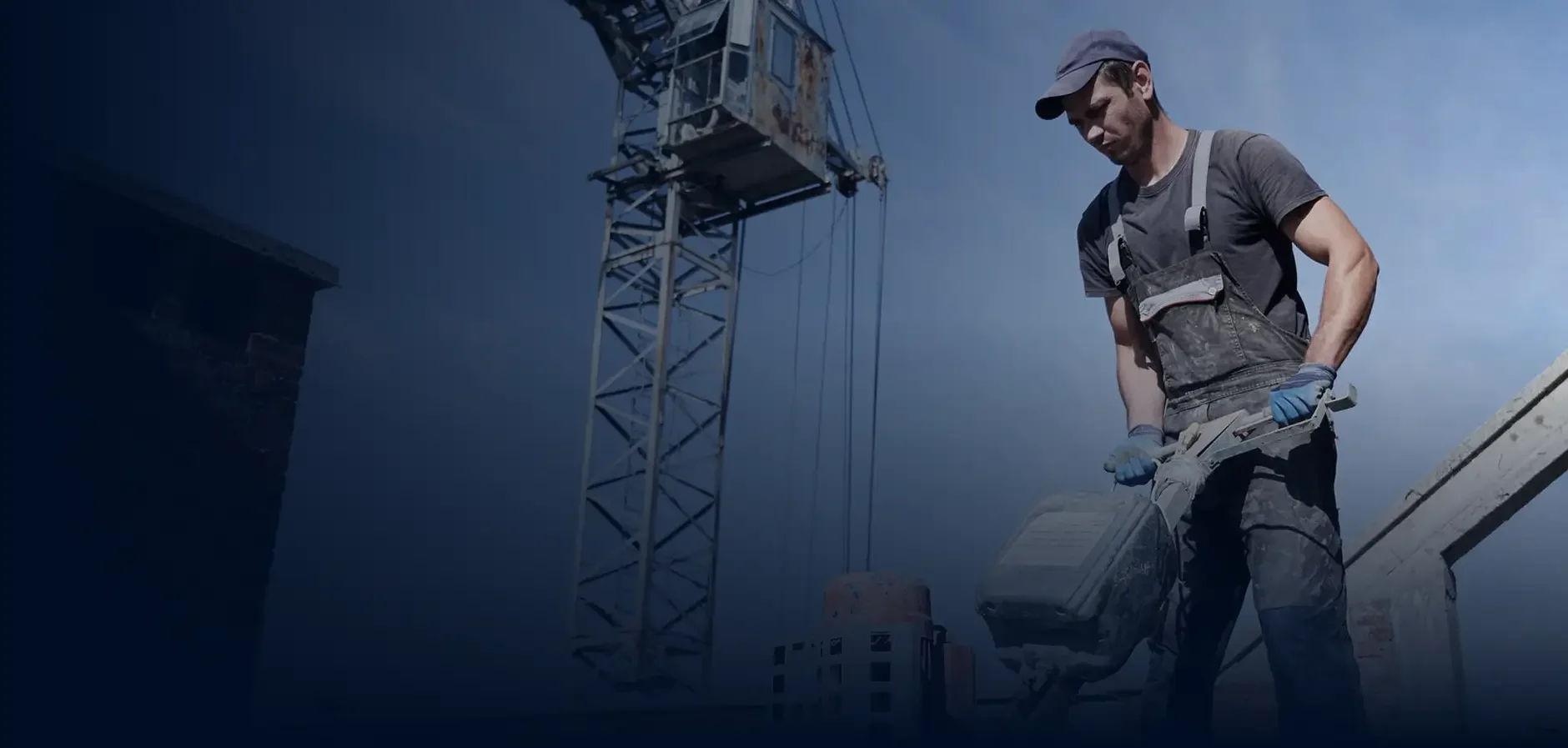 A construction worker wearing a cap, gloves, and dark work clothes, holding a power tool at a construction site with a crane in the background.