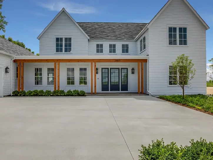 Front view of a modern white house with a large driveway, black front doors, and a wooden pergola over the entrance, with landscaping and a small tree on the right.
