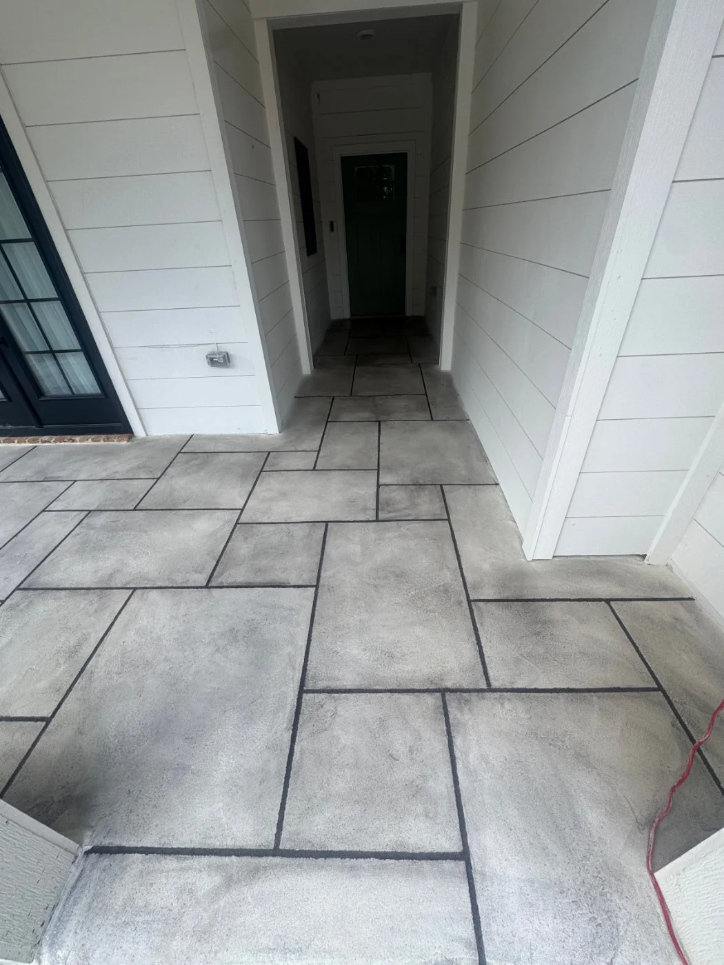 Exterior porch area with large concrete tiles, white painted siding, and a black door with glass panes. There is a small gap between the porch and the door leading into a dark interior space.