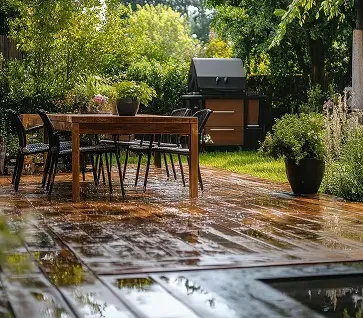 An outdoor backyard patio with a wooden dining table, black chairs, and a barbecue grill, surrounded by greenery and potted plants.