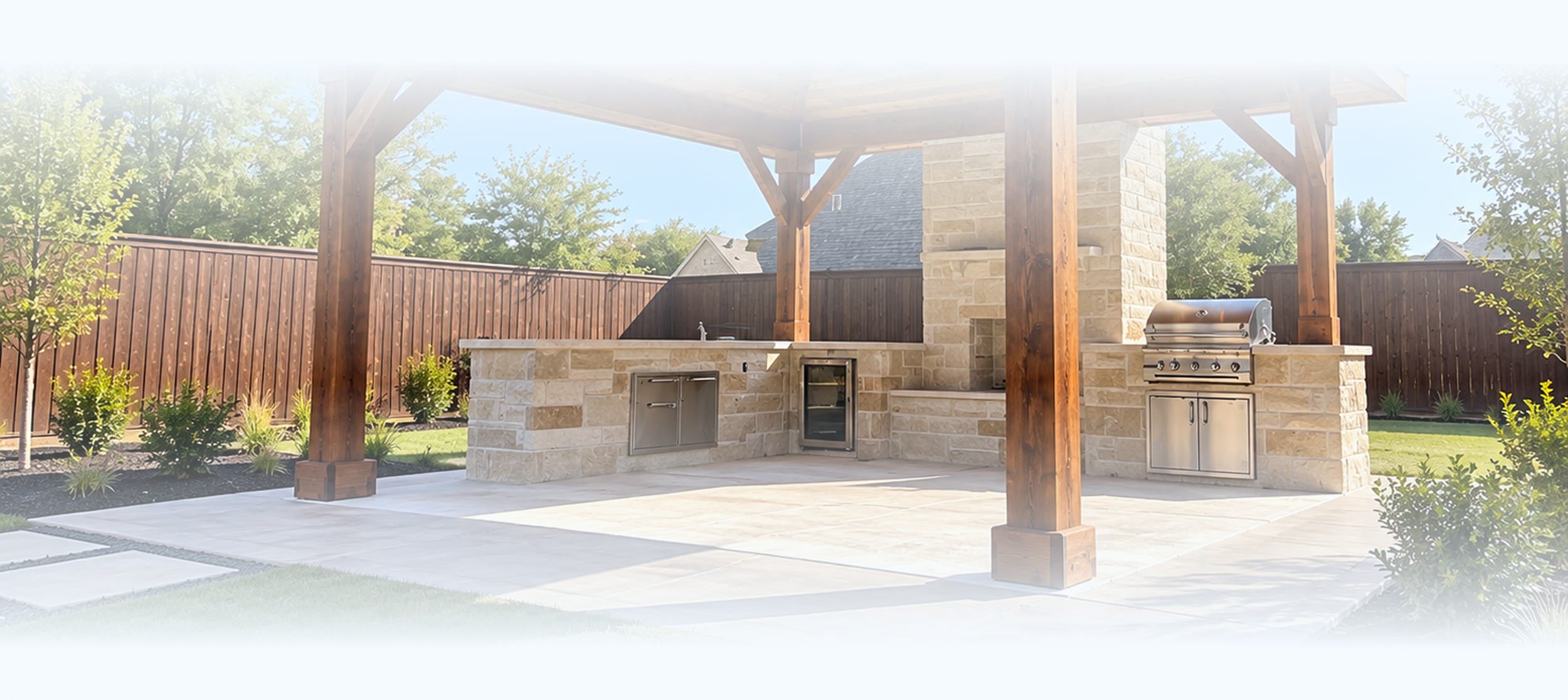 Outdoor kitchen area with stone counter, stainless steel grill, small sink, and cabinetry under a wooden pergola, surrounded by a fenced yard with trees and plants.