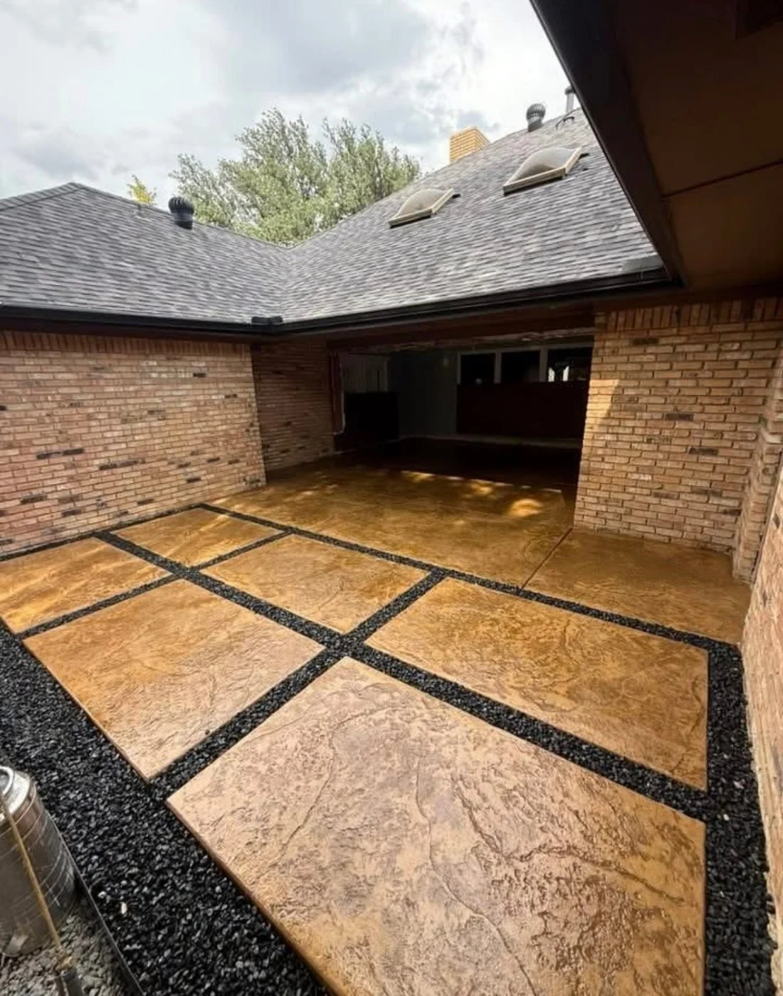 Decorative outdoor patio with large tan stones separated by black gravel borders, surrounded by brick walls and a sloped shingled roof with skylights.